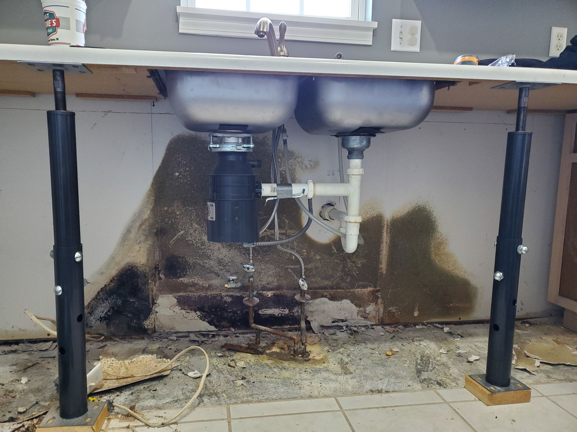 Kitchen sink with a garbage disposal supported by two black jack posts, facing a damaged, water-stained wall.