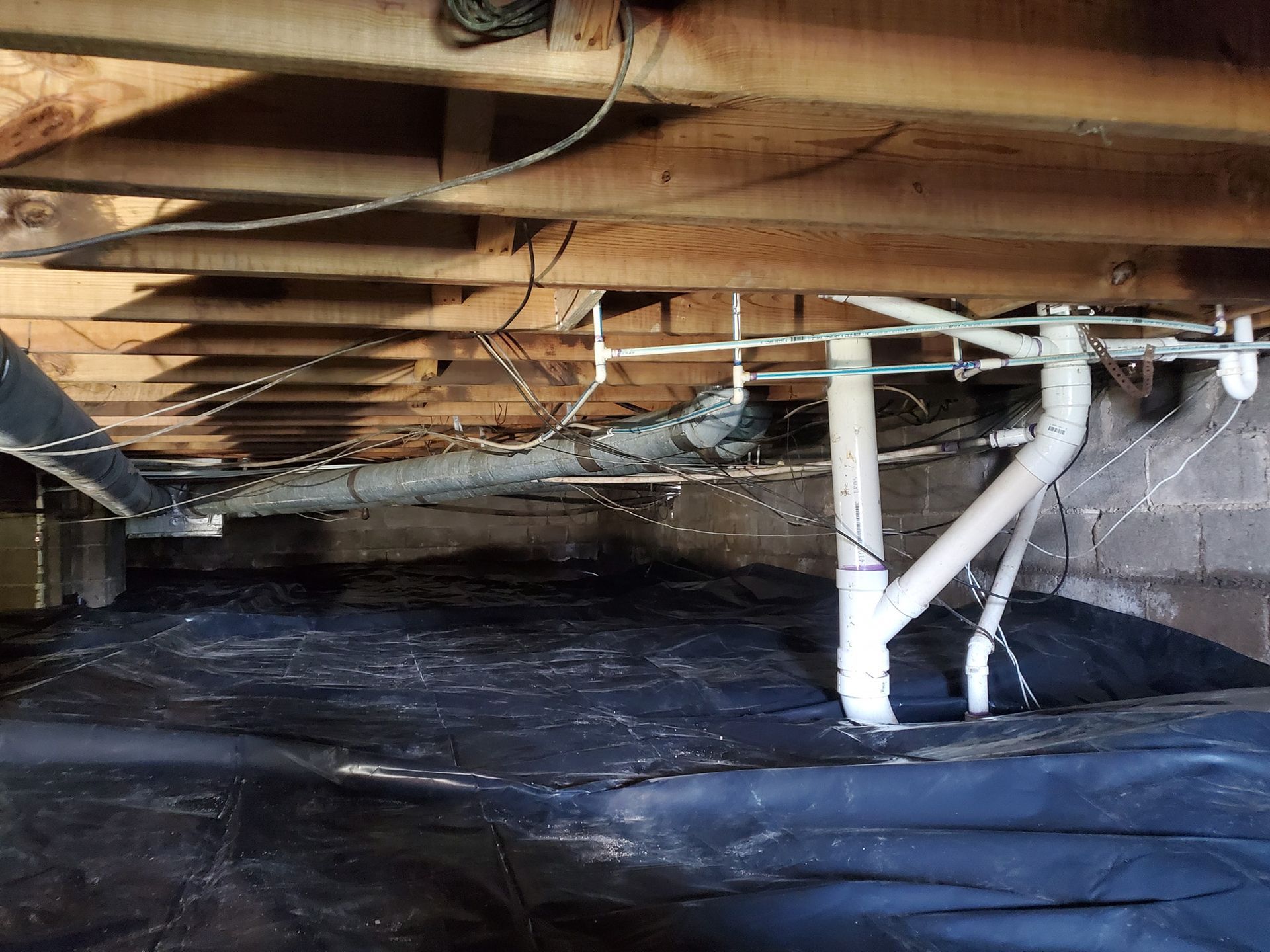Crawl space under a house floor with wooden joists, white PVC drainage pipes, and a black plastic vapor barrier.