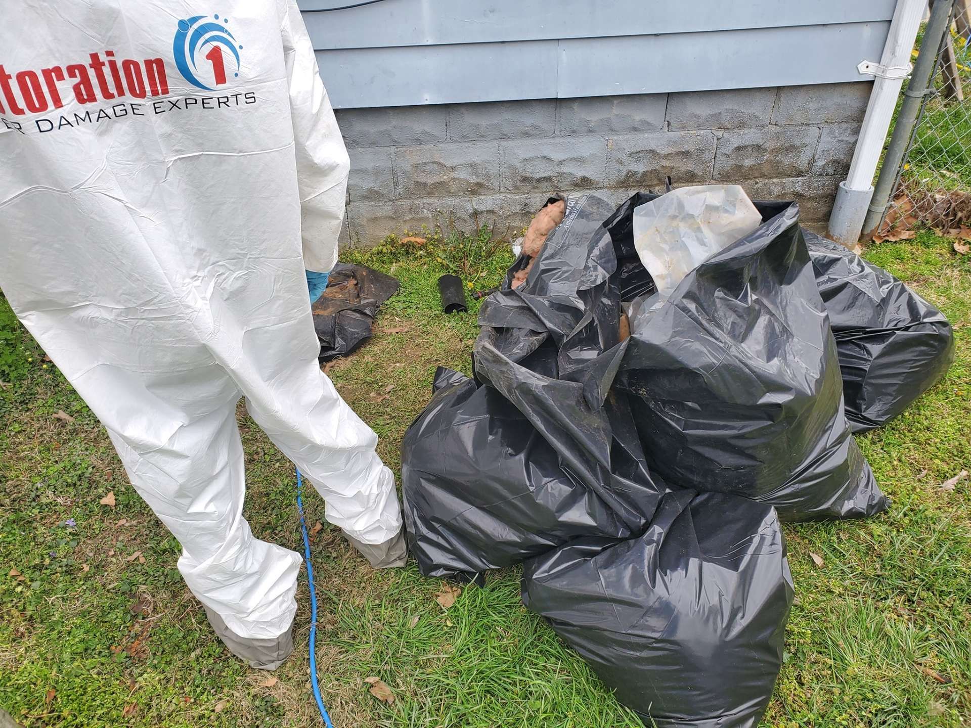 A person in protective gear stands beside a pile of black garbage bags on the grass near a building.