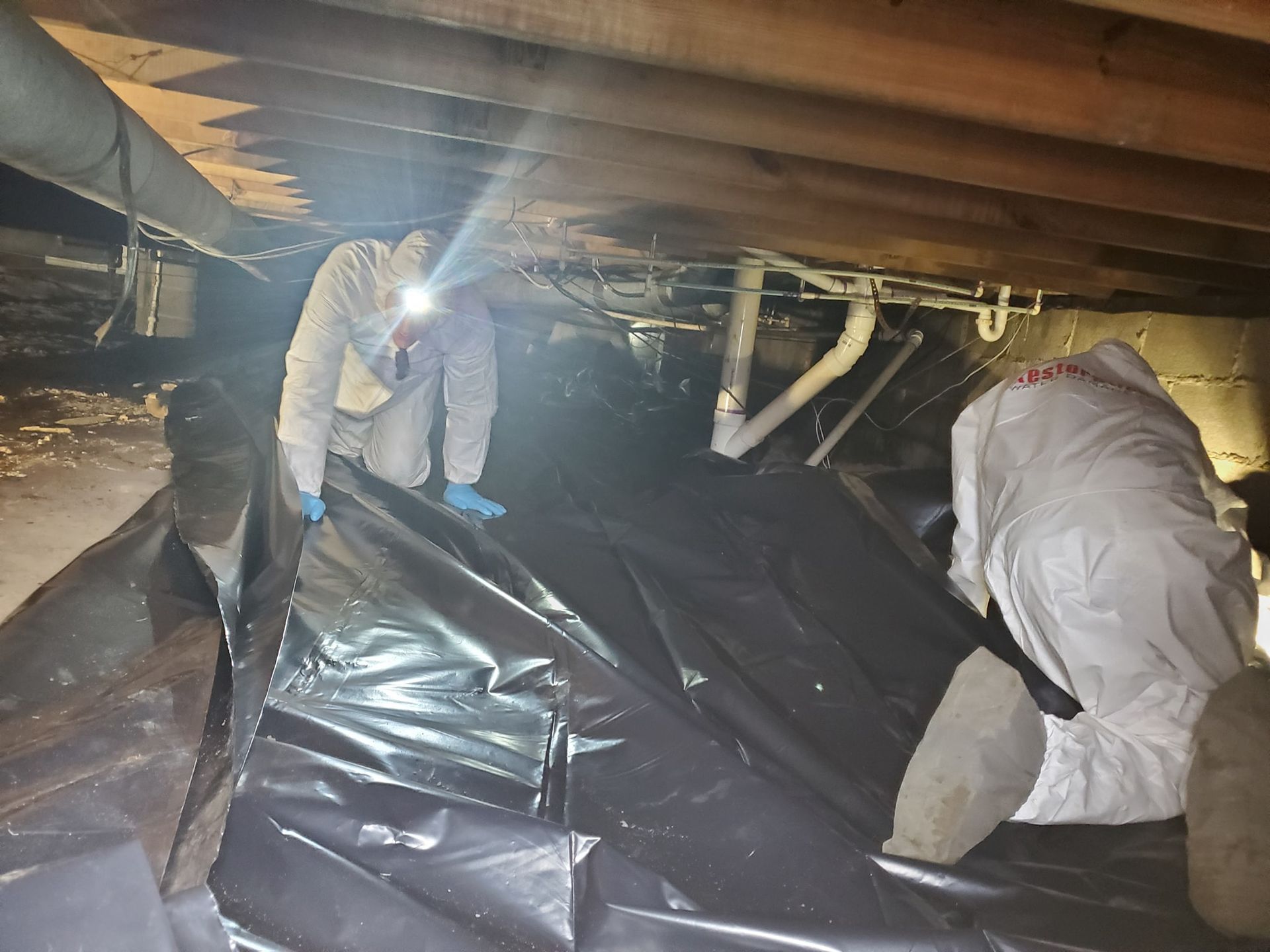 Two people in white protective suits work in a dark crawl space, installing black plastic sheeting over the ground.