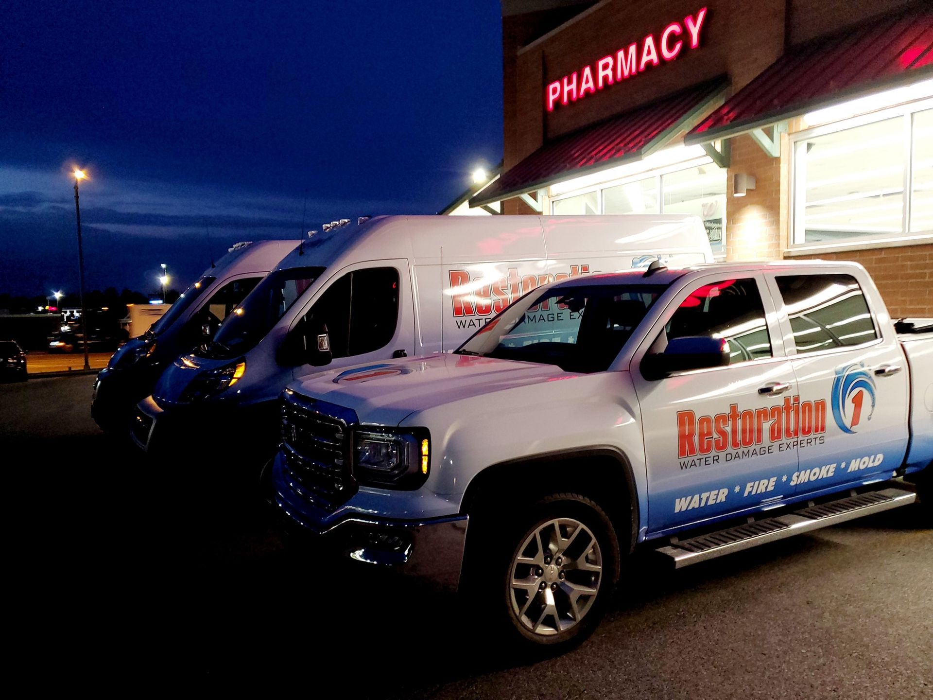 White Restoration 1 work trucks and a van parked in front of a pharmacy at twilight.