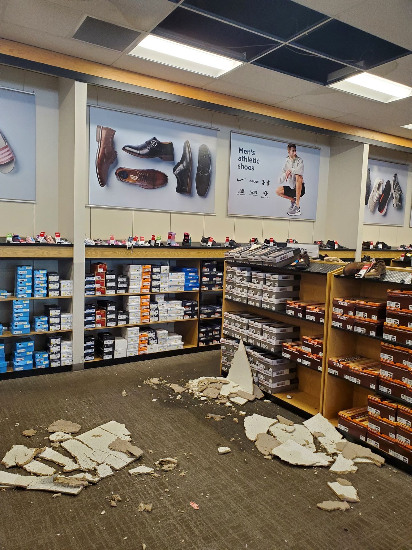A shoe store aisle with collapsed ceiling tiles and debris scattered across the carpeted floor.