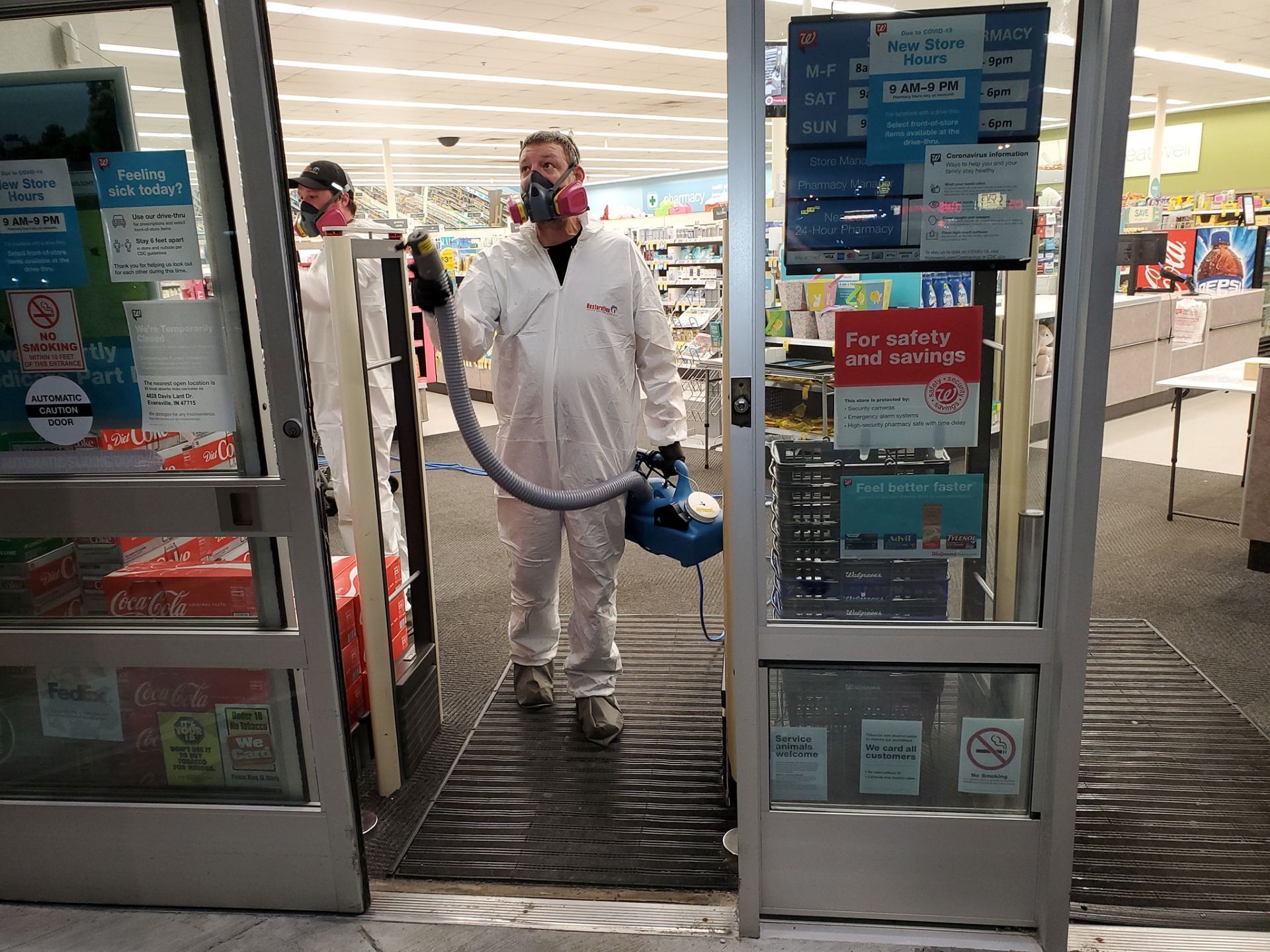 Two people in white protective suits and masks use a handheld disinfectant sprayer while entering a store doorway.