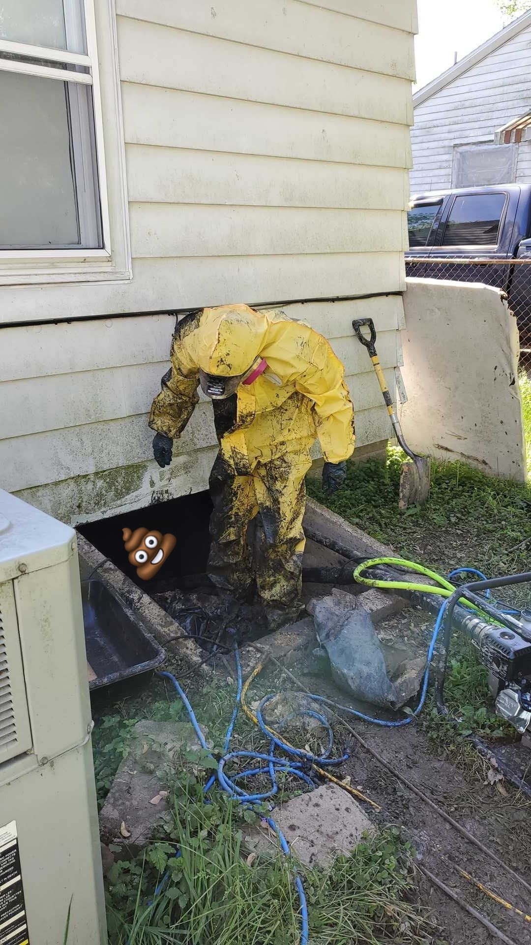 A person in a yellow hazmat suit stands in a dark crawl space under a building, reaching toward a large cartoon poop icon.