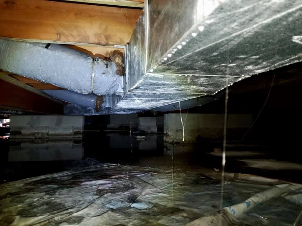 Water drips from a metal HVAC duct onto a dark, flooded crawl space floor beneath wooden floor joists.