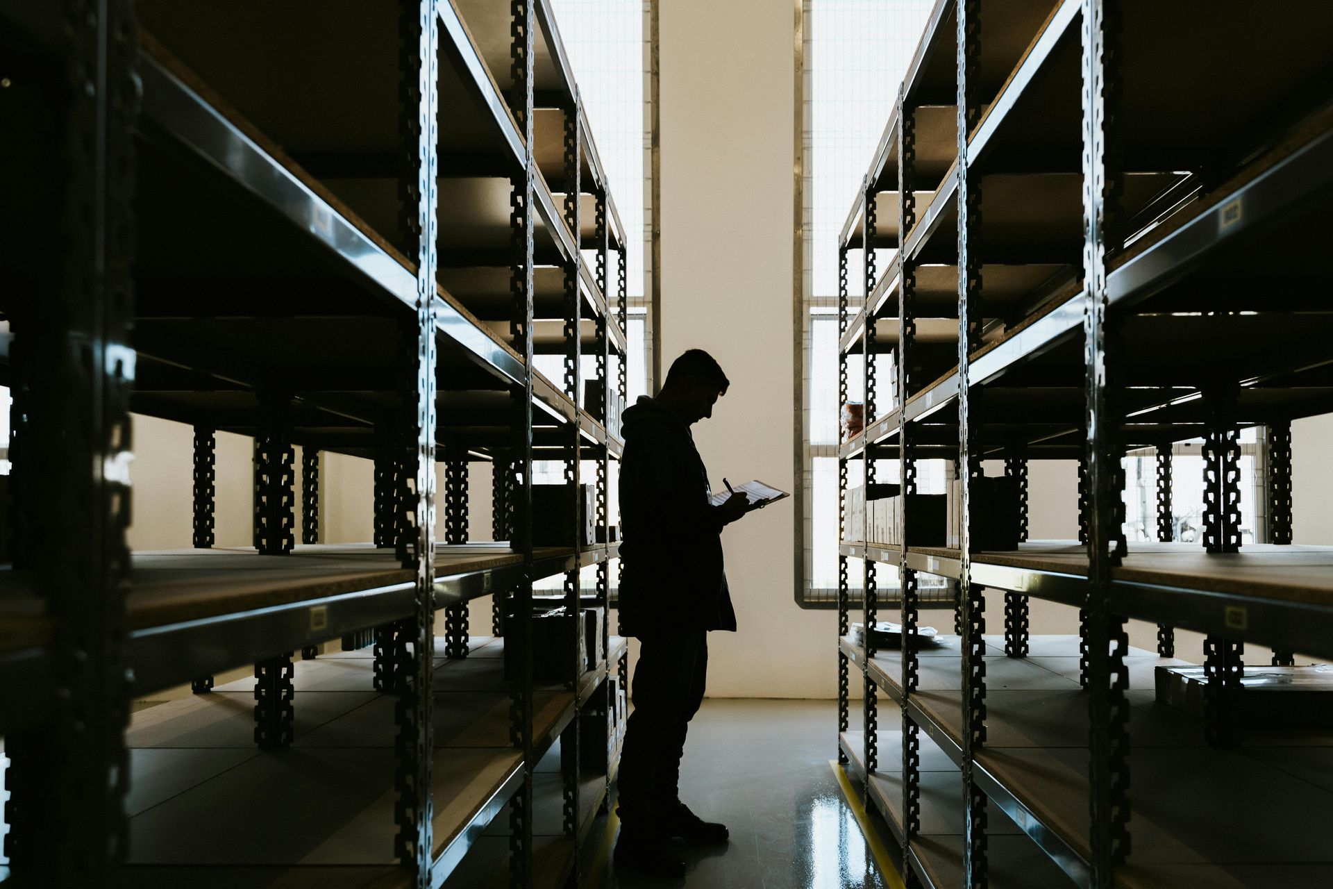 A silhouette of a person standing between tall, empty warehouse shelving units, checking a handheld device.