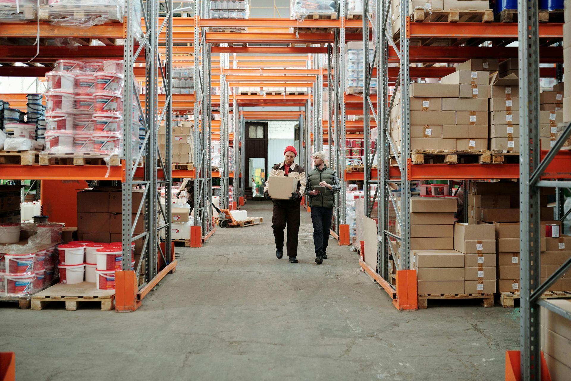 Two people walk through a warehouse aisle lined with tall orange industrial shelves stacked with goods and cardboard boxes.