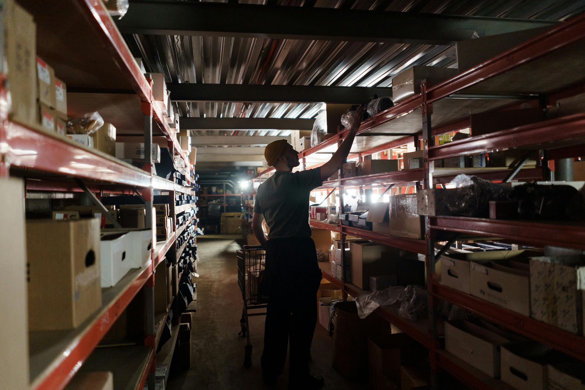 A person reaches for an item on a shelf in a dimly lit warehouse filled with rows of metal shelving and storage boxes.
