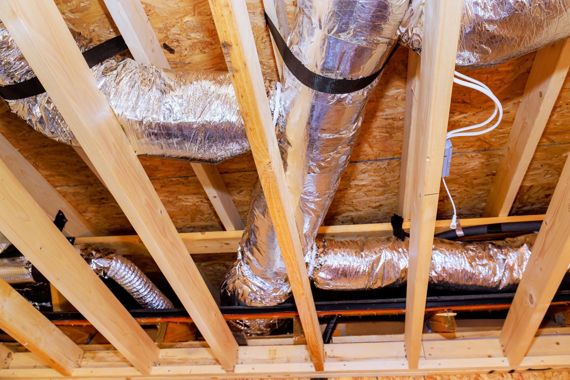 Foil-wrapped HVAC air ducts suspended with black straps between wooden roof joists.