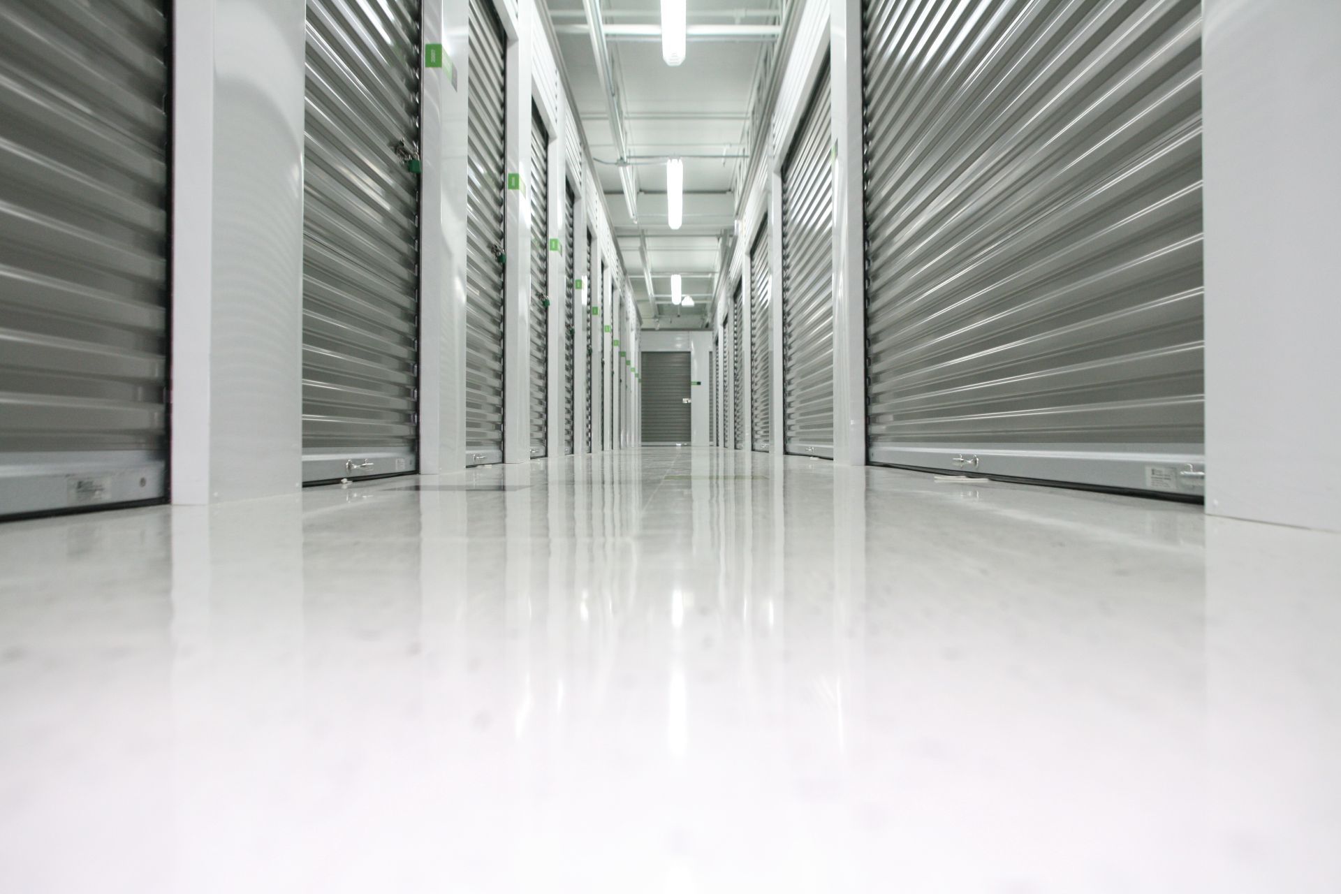 A narrow, well-lit hallway of a self-storage facility featuring rows of closed metal rolling doors and shiny white floors.