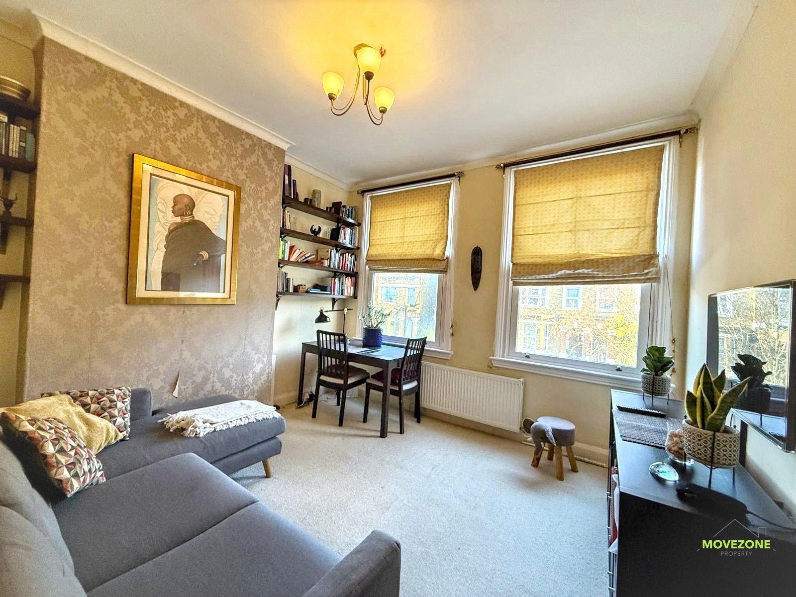 Bright living room with gray sofa, small dining table, tan blinds, and potted plant by the window.