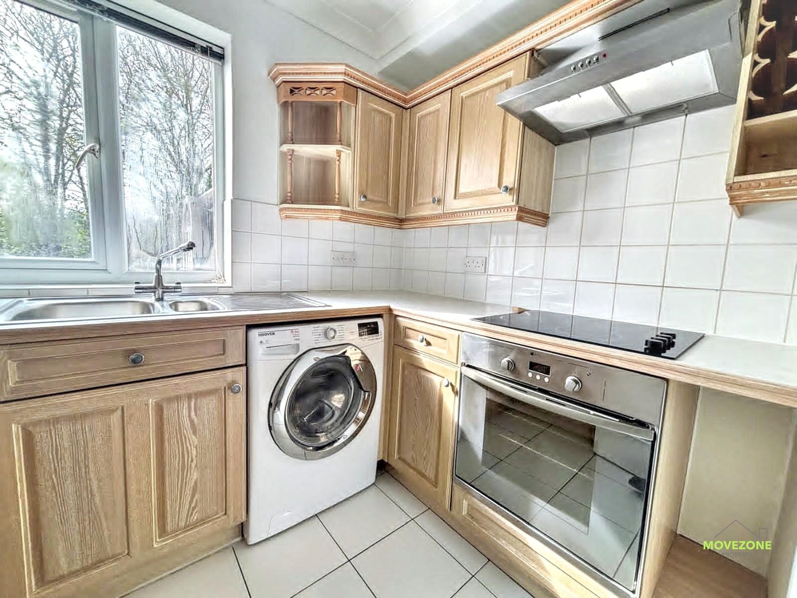 Small corner kitchen with light wood cabinets, white tile, a window, and a washing machine under the counter.