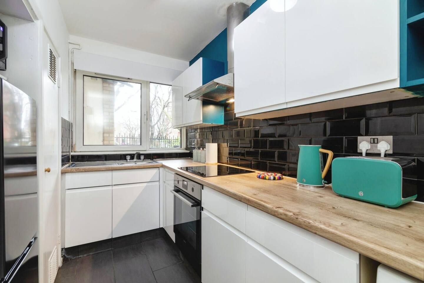 Modern white kitchen with wood countertops, black tile backsplash, and teal appliances by a window
