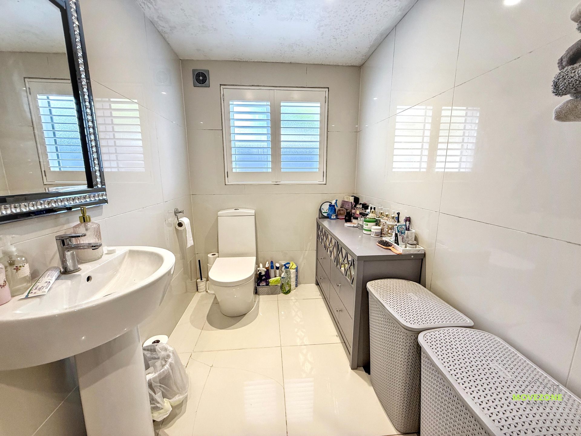 Bright white bathroom with sink, toilet, window, mirror, and wicker laundry bins.