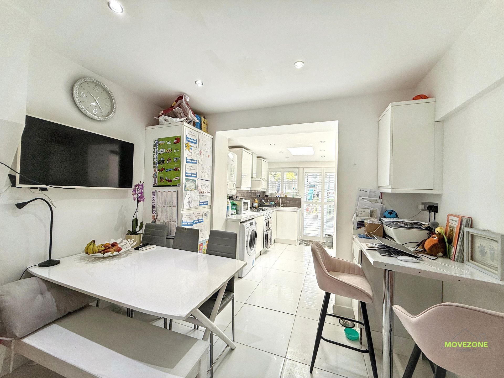Bright white kitchen with a table, wall-mounted TV, bar stools, and an open doorway to a sunlit utility area