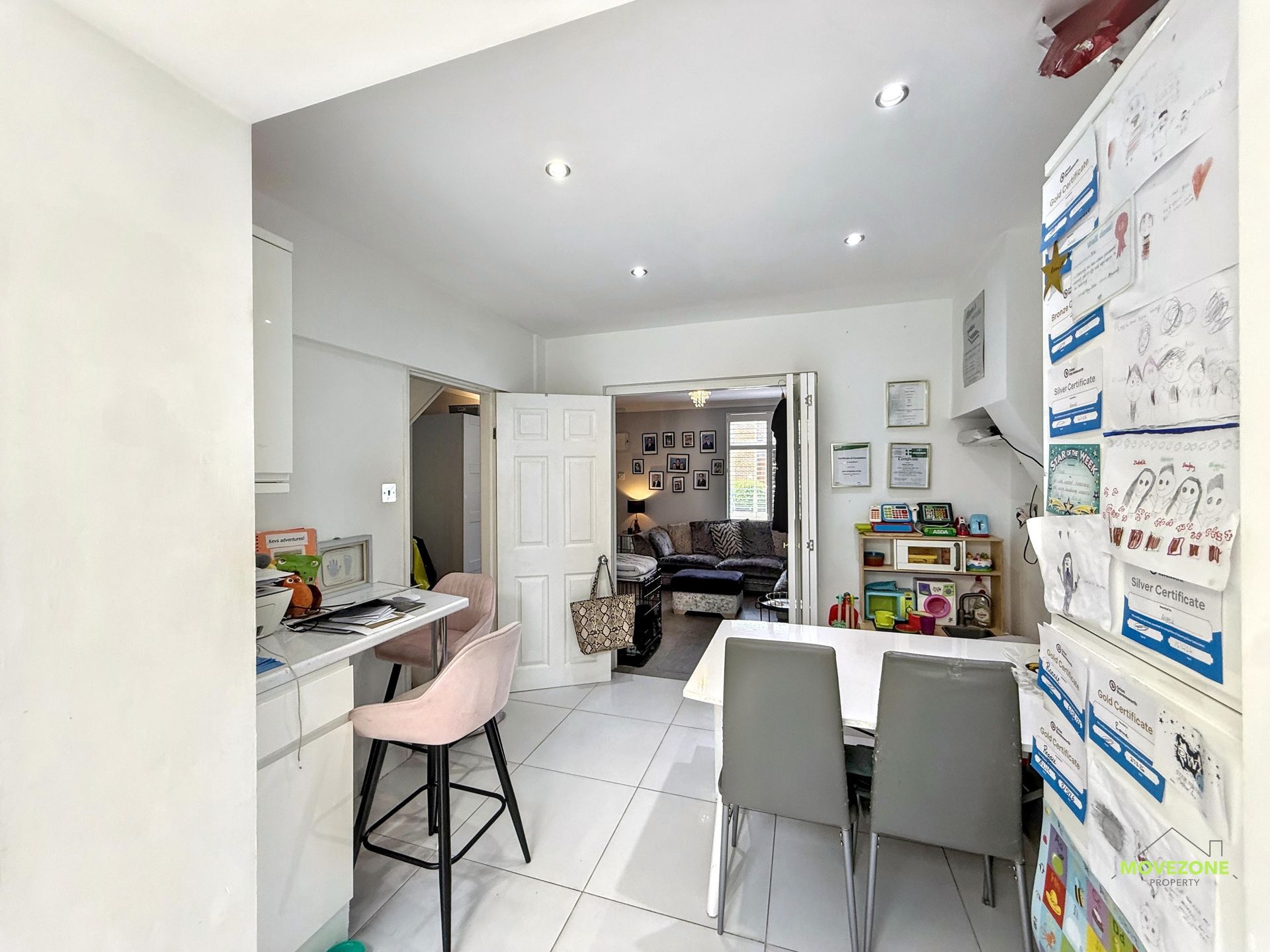 Bright kitchen-dining room with white tile floors, pink stool, gray chairs, and a doorway to a living area