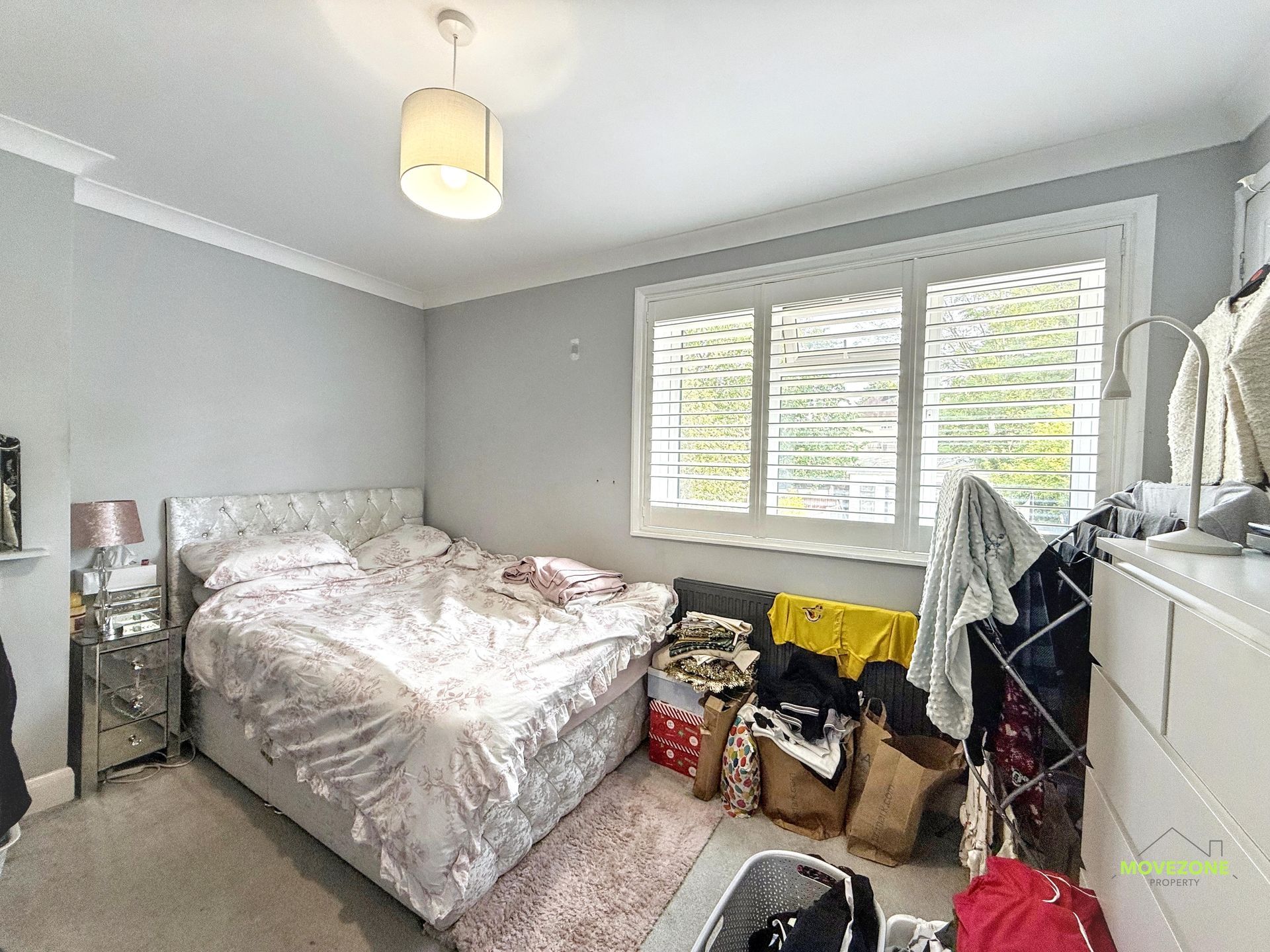 Small gray bedroom with an unmade bed, window blinds, and cluttered bags and clothes by the wall
