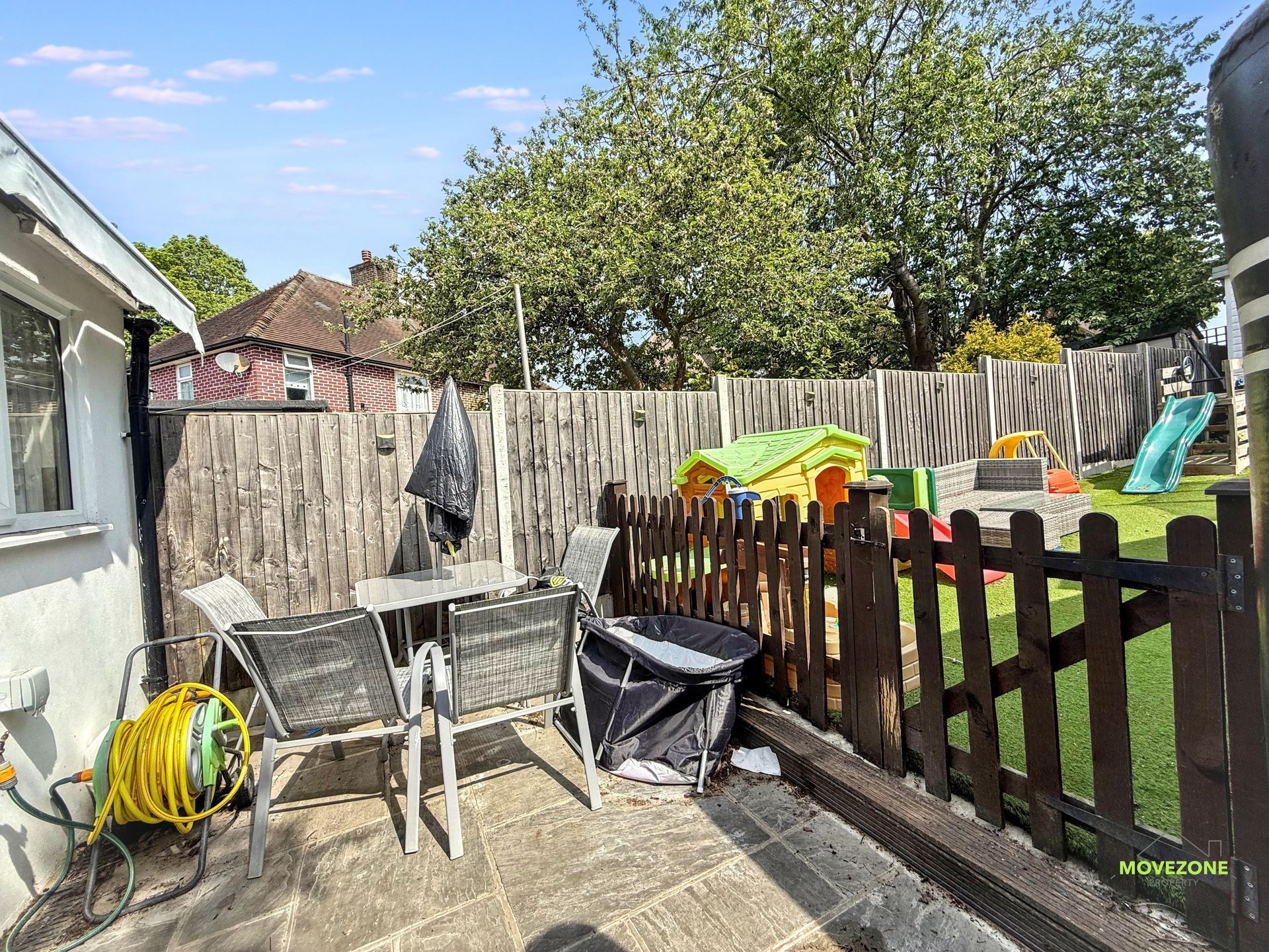 Backyard patio with chairs, table, hose reel, fence, and colorful kids’ slide under a sunny sky
