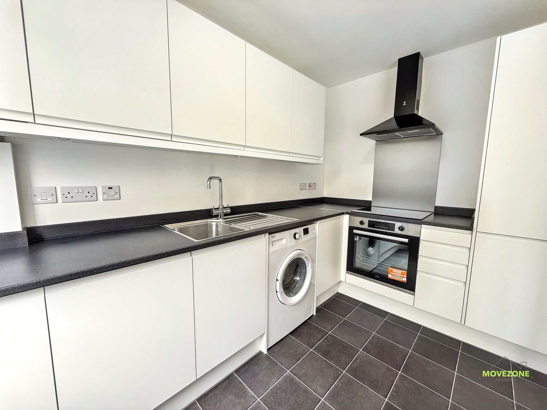 Modern white kitchen with black countertops, stainless stove, washing machine, and dark tiled floor