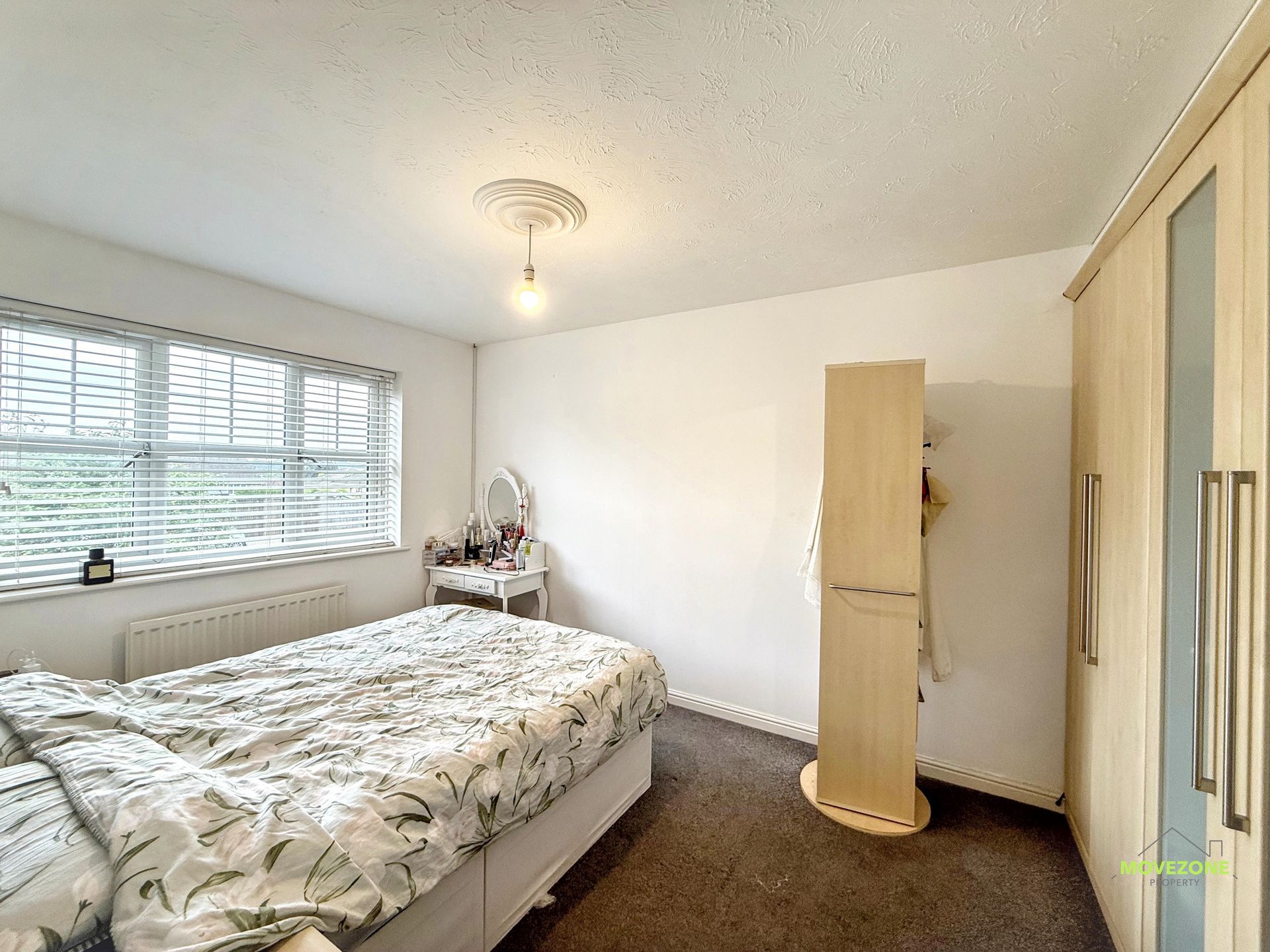 Bright bedroom with a bed, window blinds, small desk, and tall wooden cabinet against a white wall
