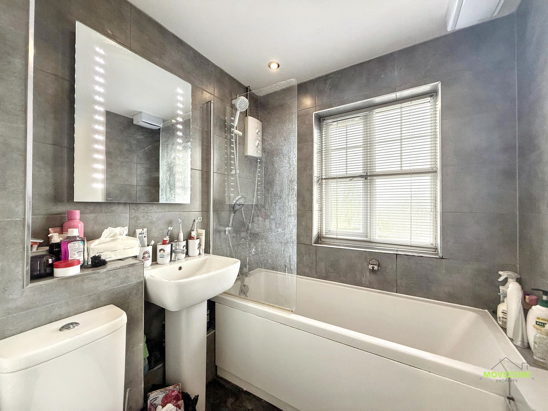 Compact bathroom with gray tiled walls, white bathtub, pedestal sink, toilet, and window with blinds.