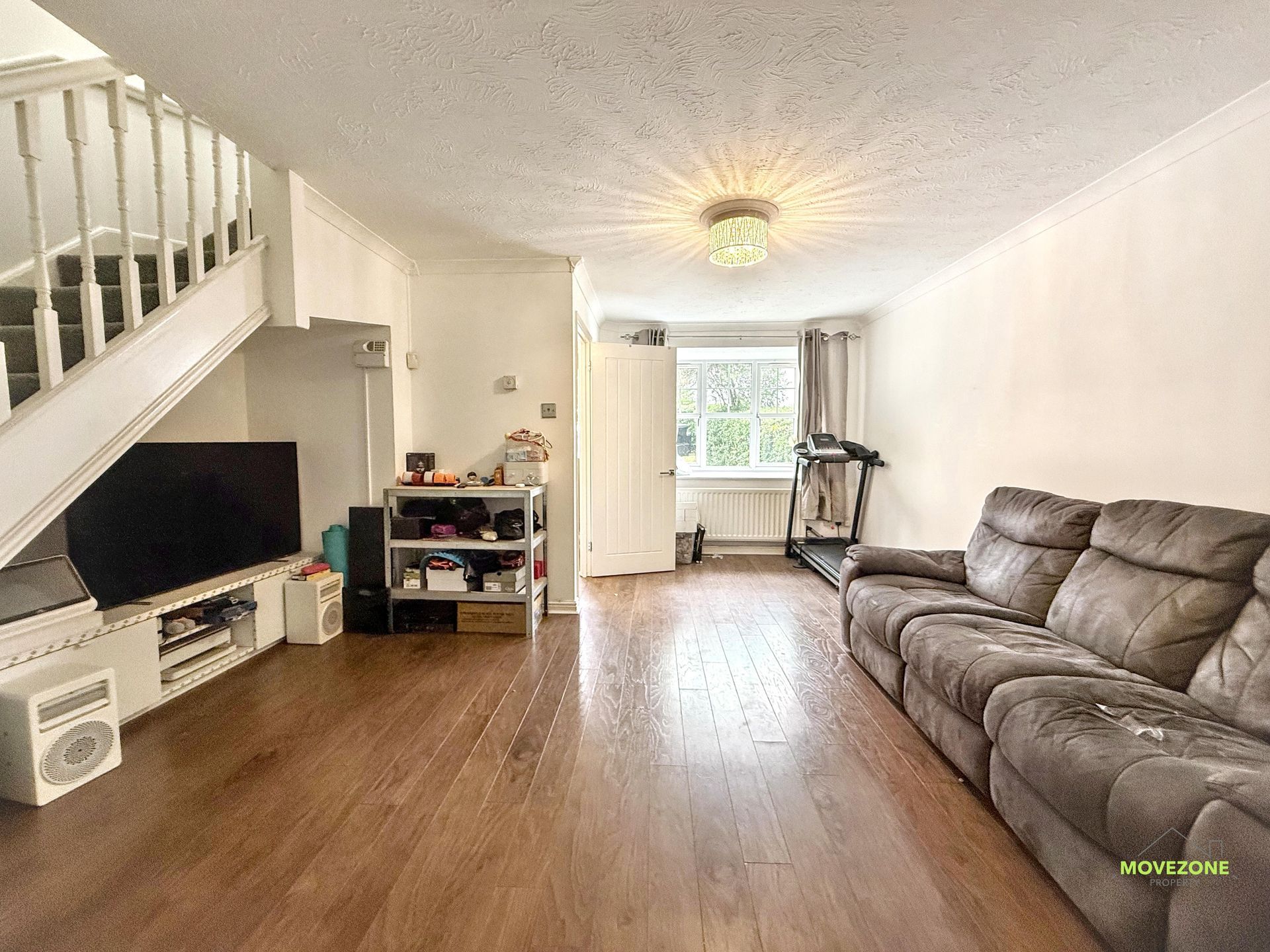 Bright living room with gray sectional, TV under stairs, wood floors, and a treadmill by the window.