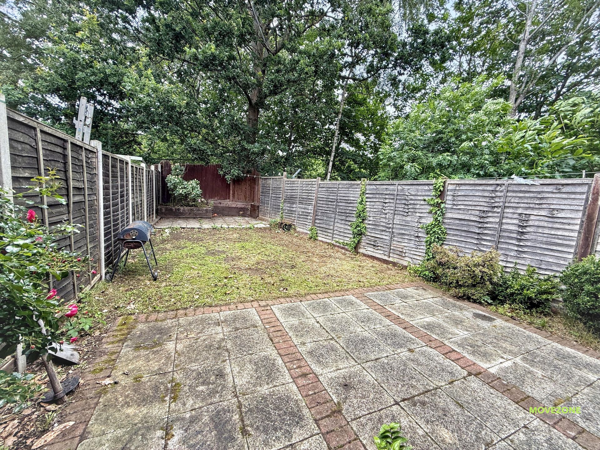 Backyard with patio tiles, grassy strip, wooden fences, shrubs, and a large tree overhead