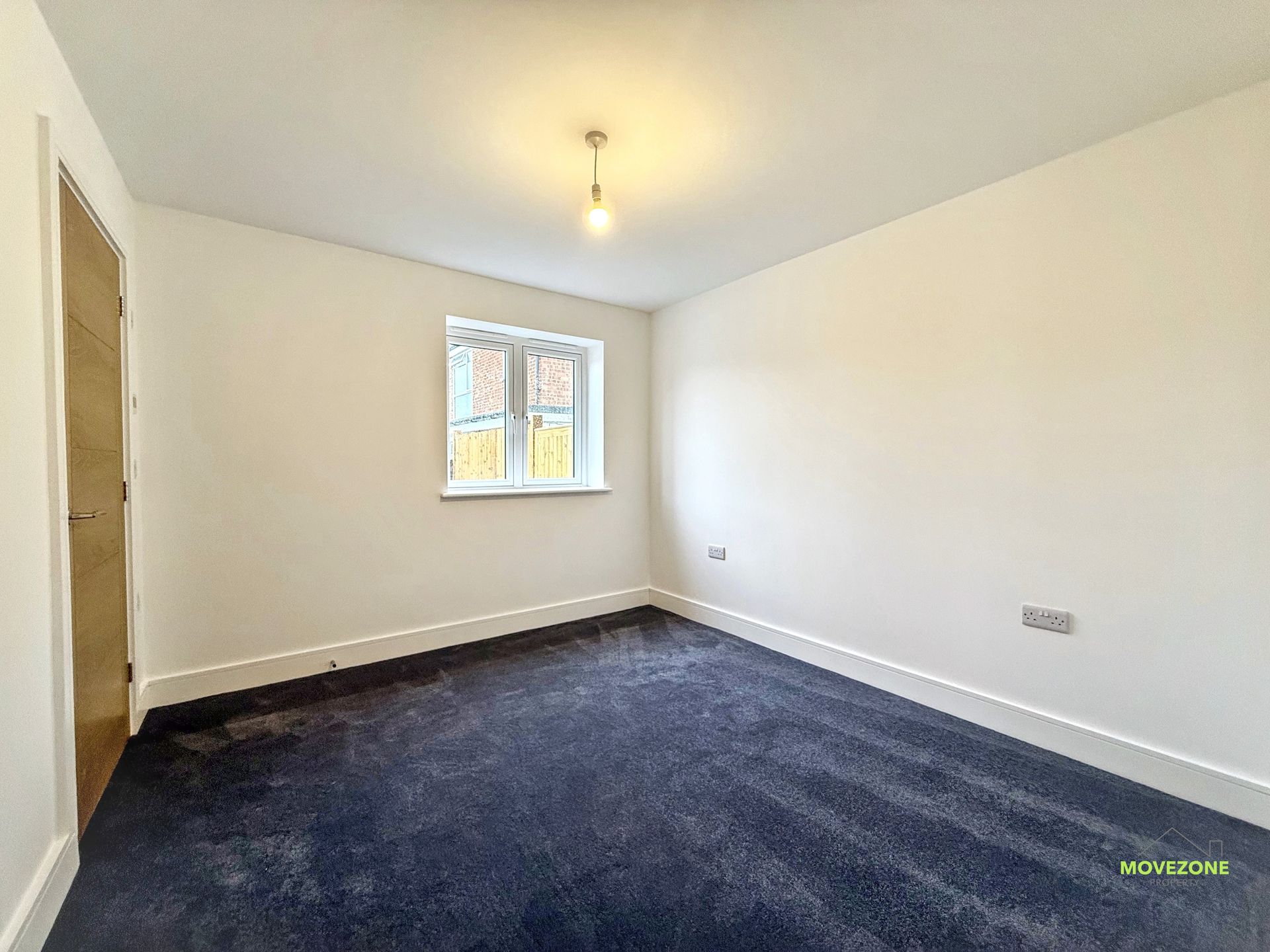Empty unfurnished room with white walls, dark carpet, window, and a single ceiling light.