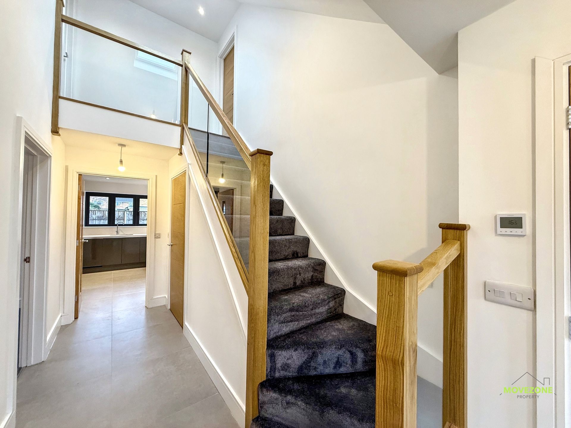Hallway with carpeted staircase, glass railing, and white walls leading to a kitchen.