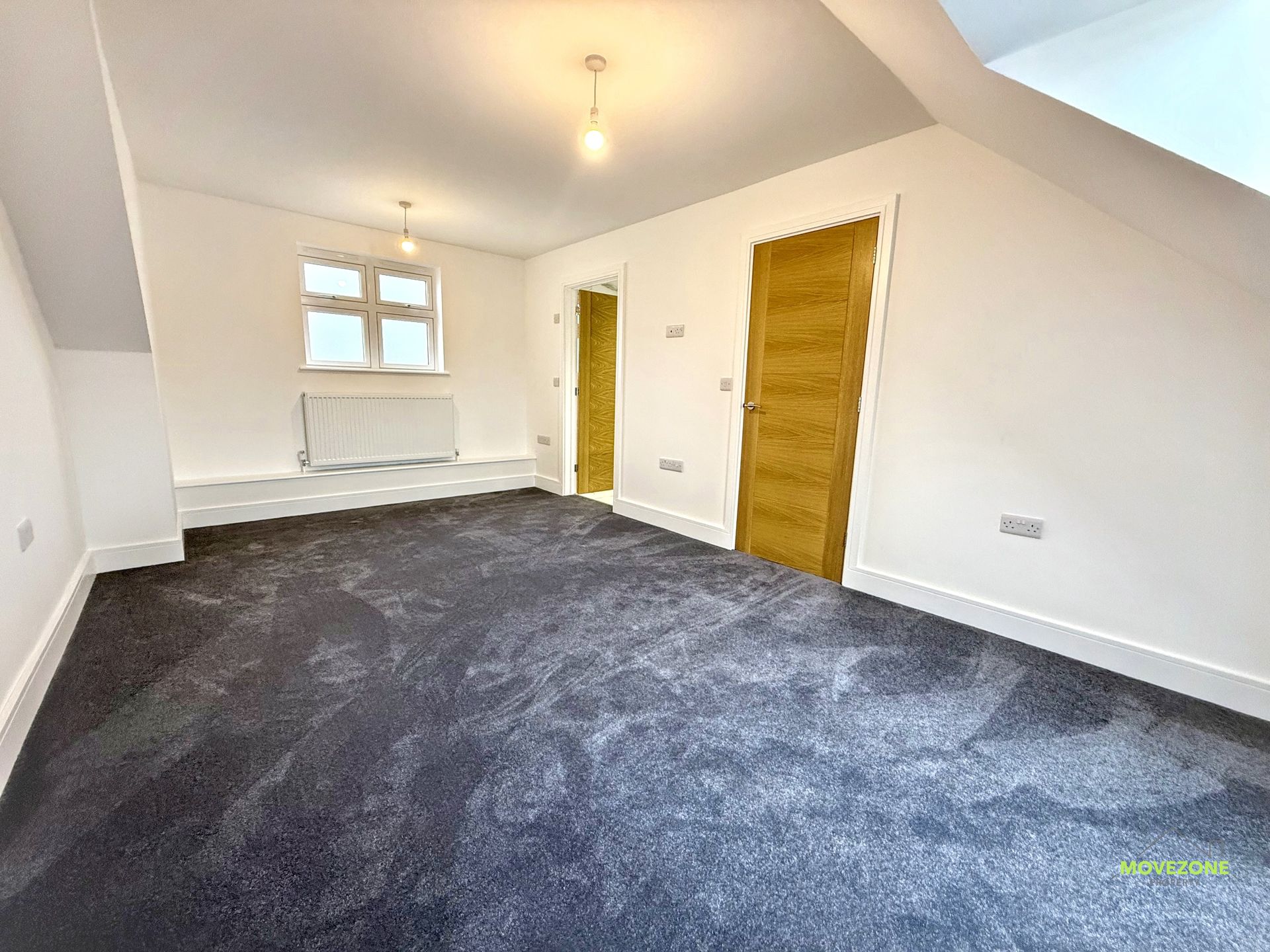 Empty attic bedroom with sloped ceiling, white walls, gray carpet, window, radiator, and two wooden doors