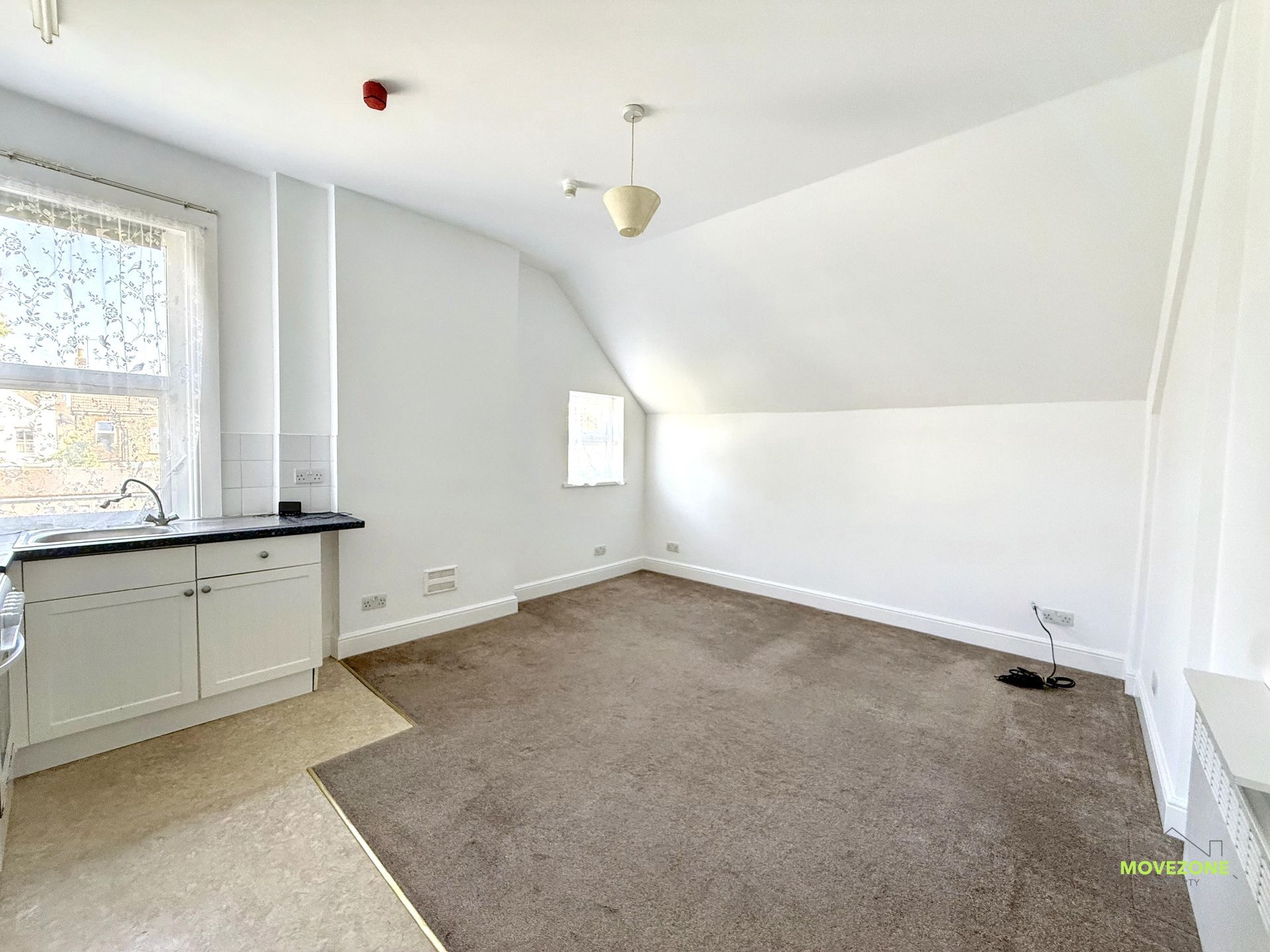 Empty bright room with beige carpet, white walls, small window, and cabinet under a sloped ceiling