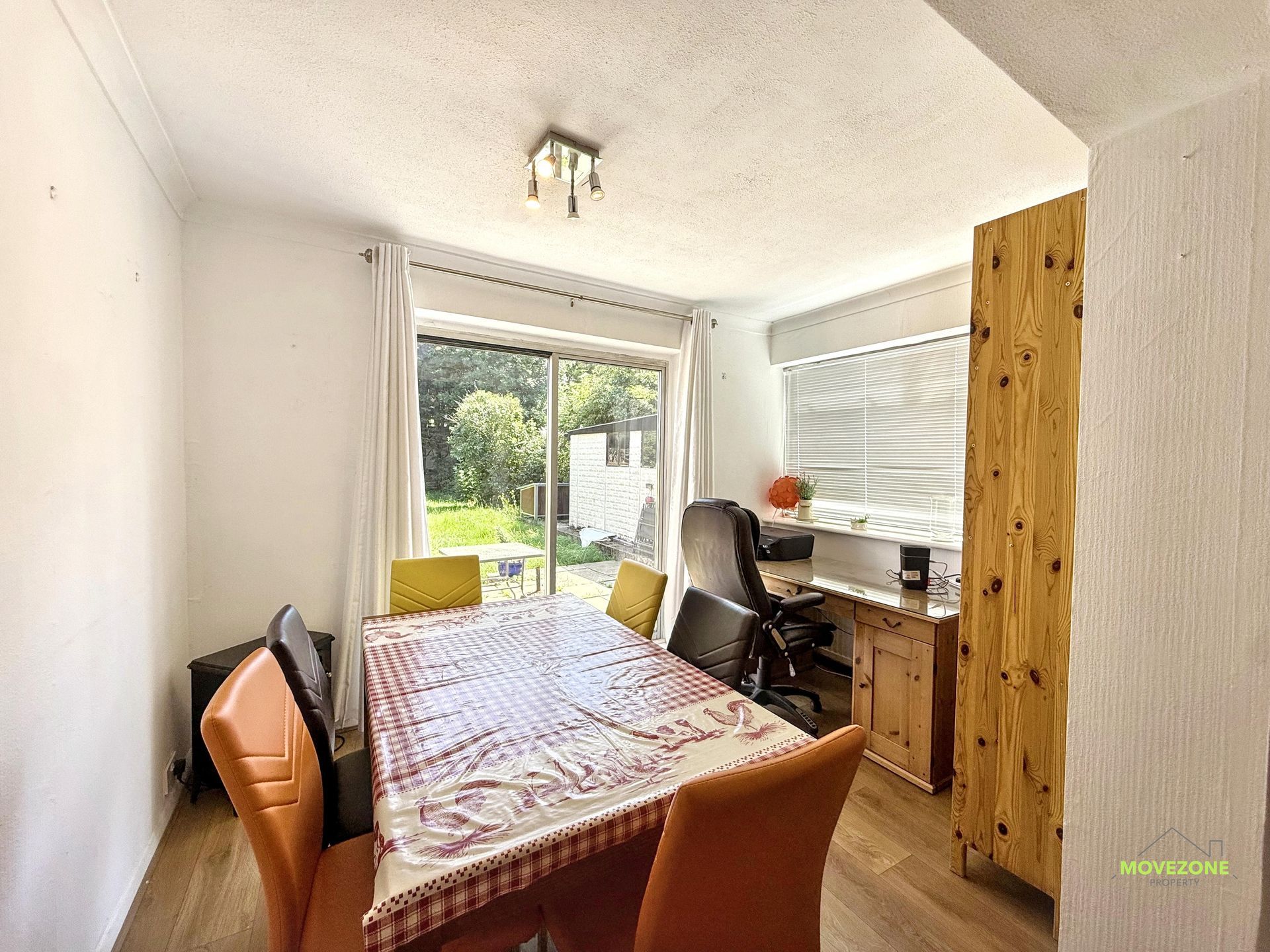 Small dining room with table, chairs, desk by window, and garden view through glass doors.