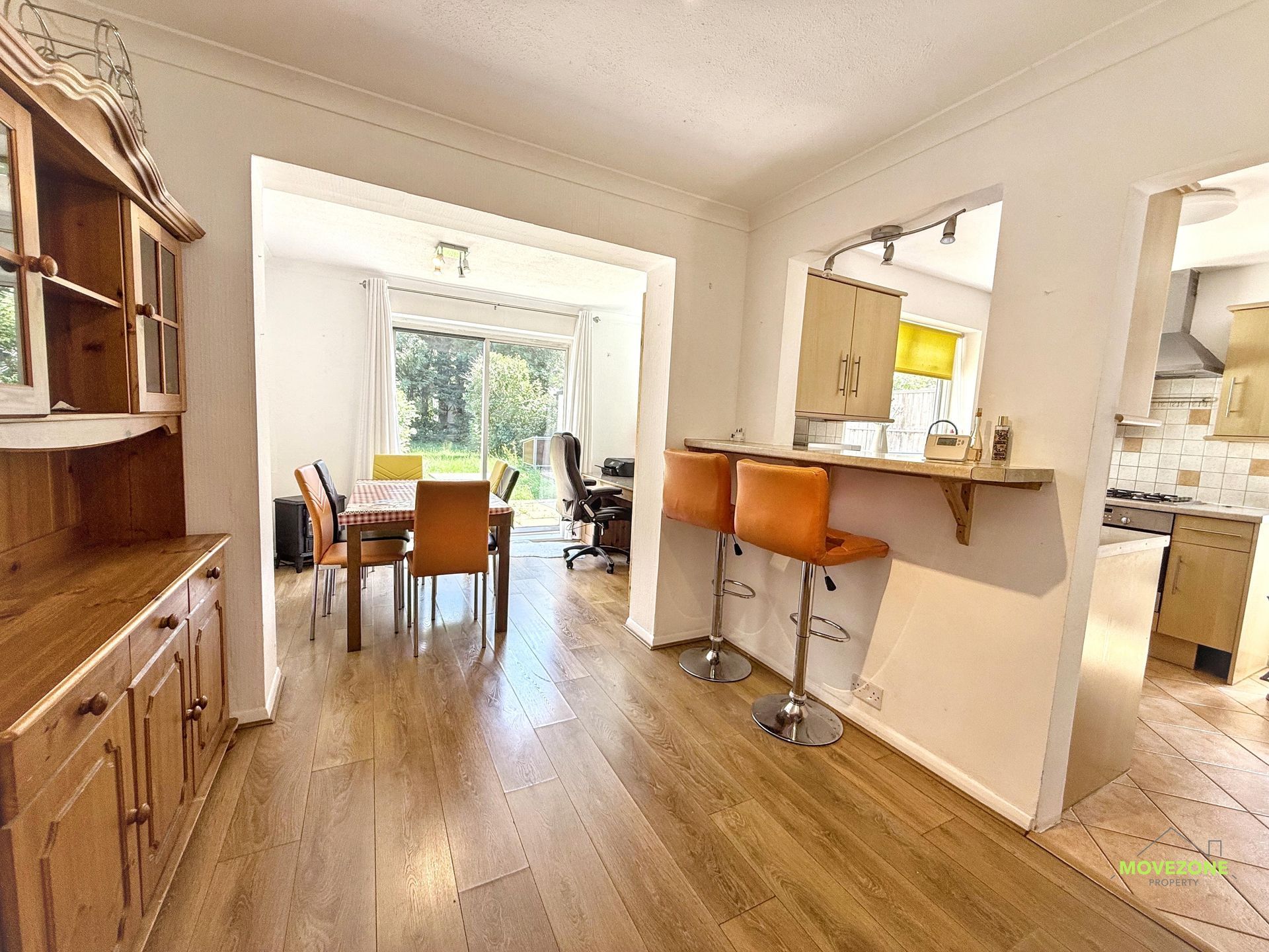 Bright open-plan dining and kitchen area with wood floors, bar stools, and a large window beyond.