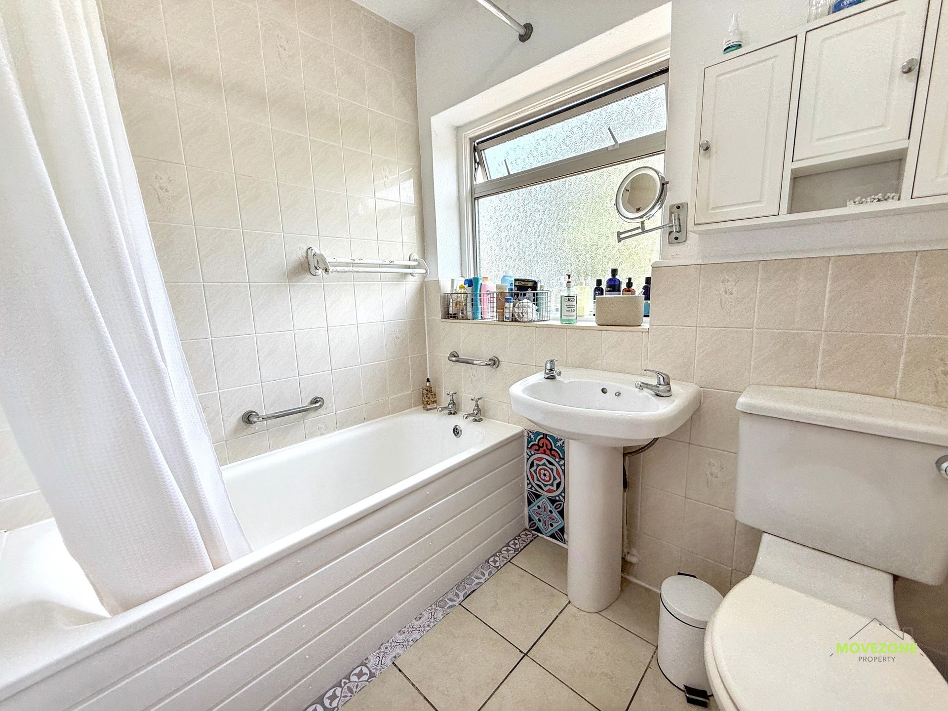 Small beige-tiled bathroom with a bathtub, pedestal sink, toilet, and window shelf.
