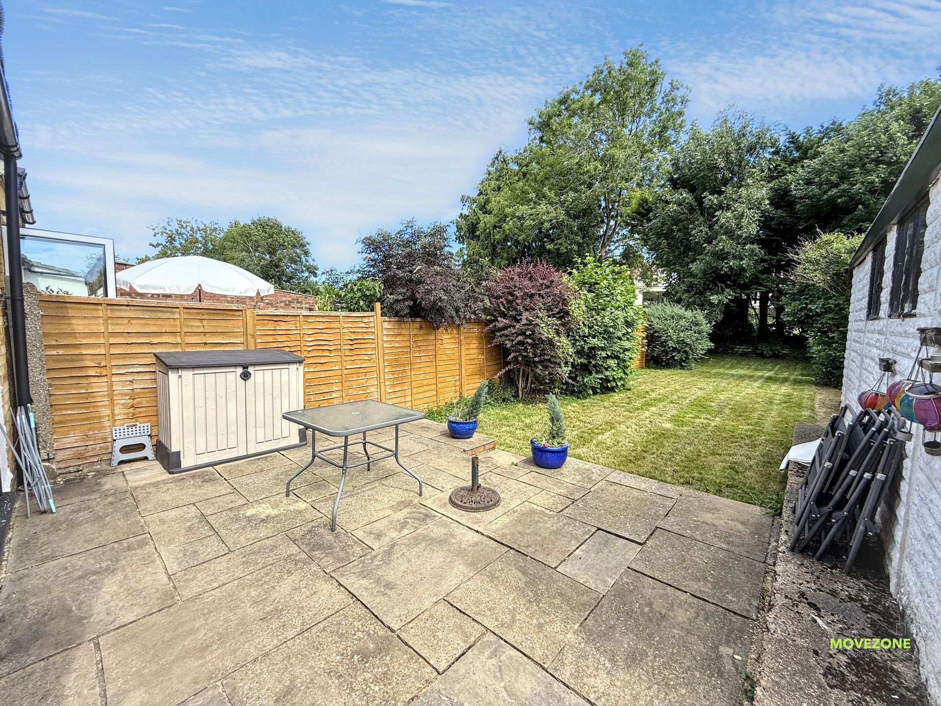 Back patio with table, storage shed, fence, potted plants, and grassy backyard under a blue sky