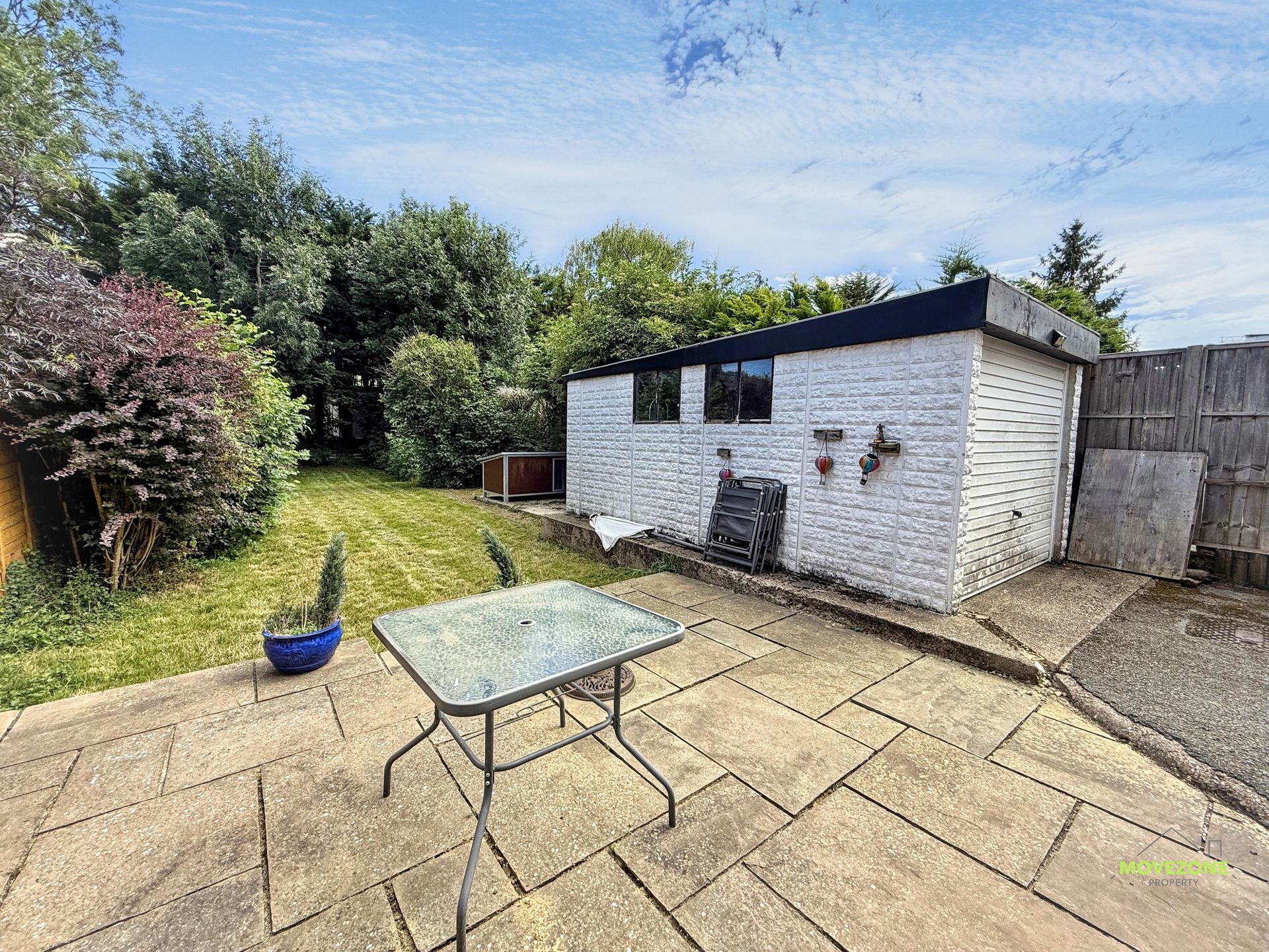 Backyard patio with a glass table, potted plant, lawn, and white shed under a blue sky