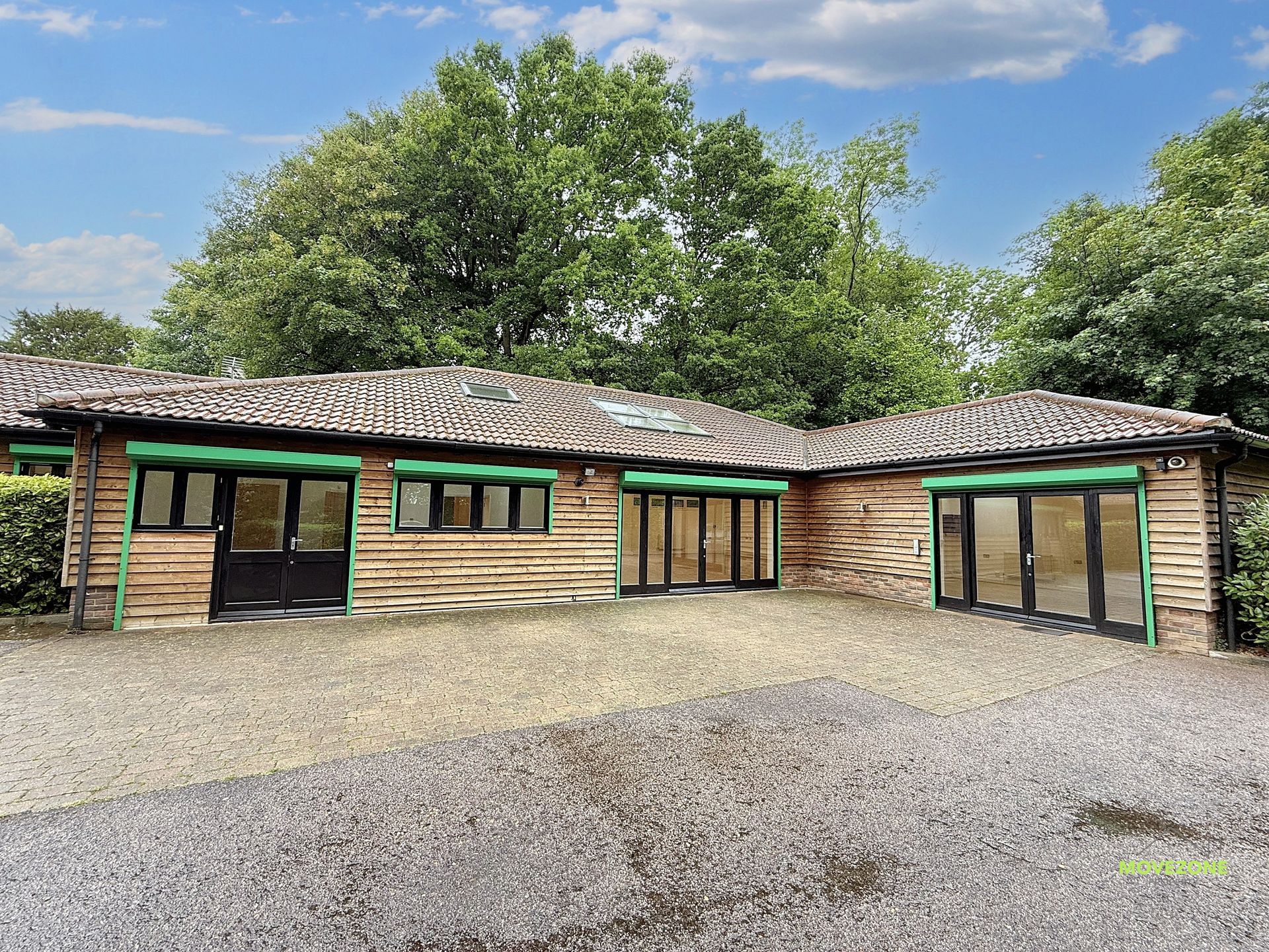 Single-story wooden building with green-trimmed windows and a sloped roof, surrounded by trees and gravel.