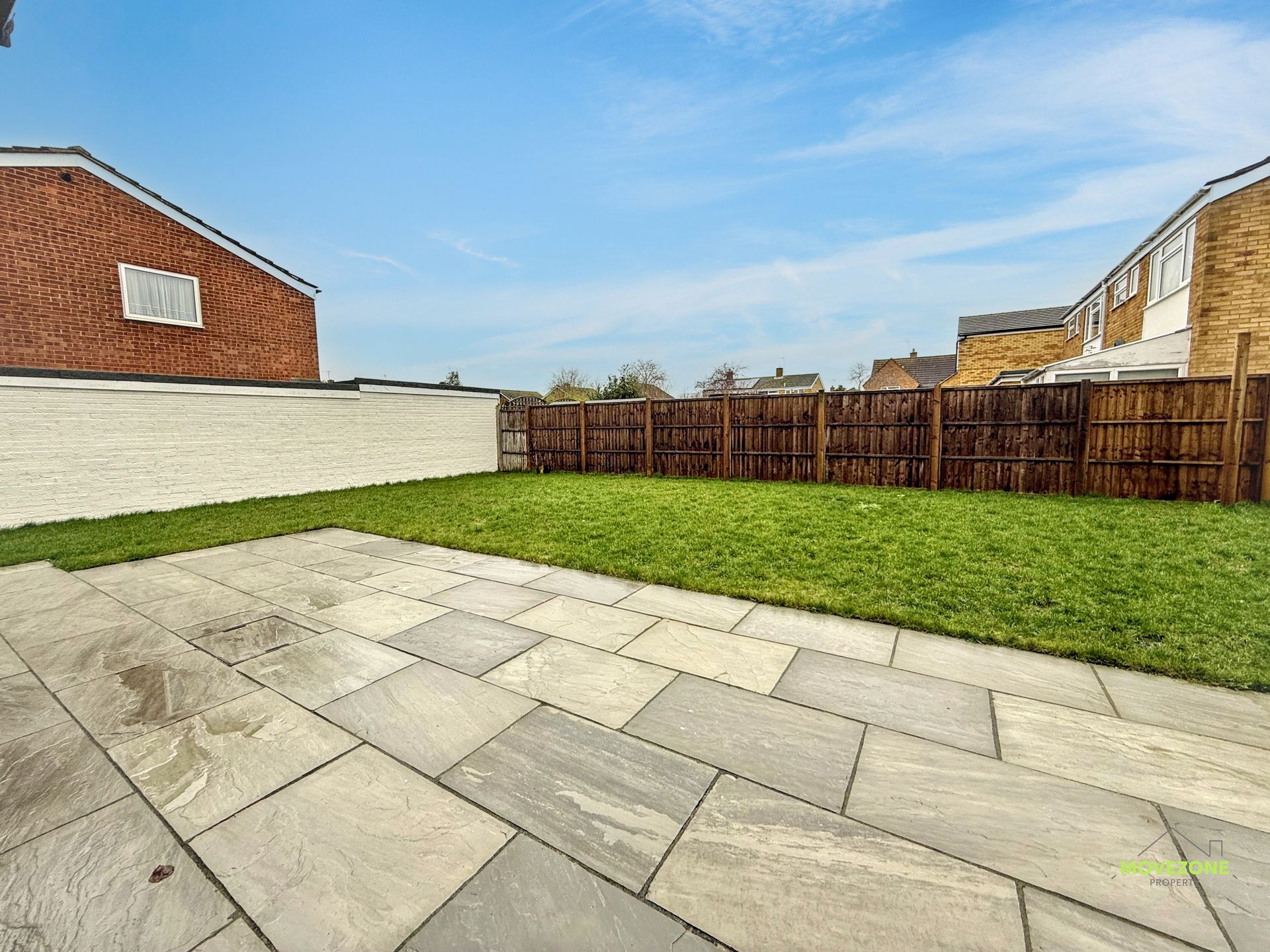 Backyard with gray stone patio, green lawn, white wall, and wooden fence under a blue sky