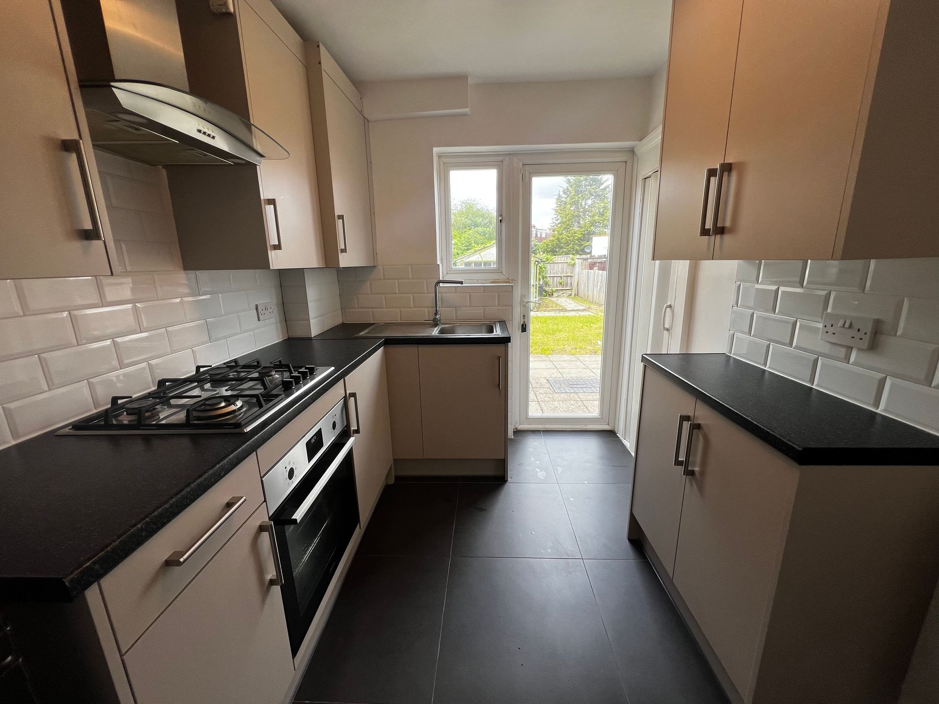 Narrow modern kitchen with beige cabinets, black countertops, white tile backsplash, and a glass door to outside