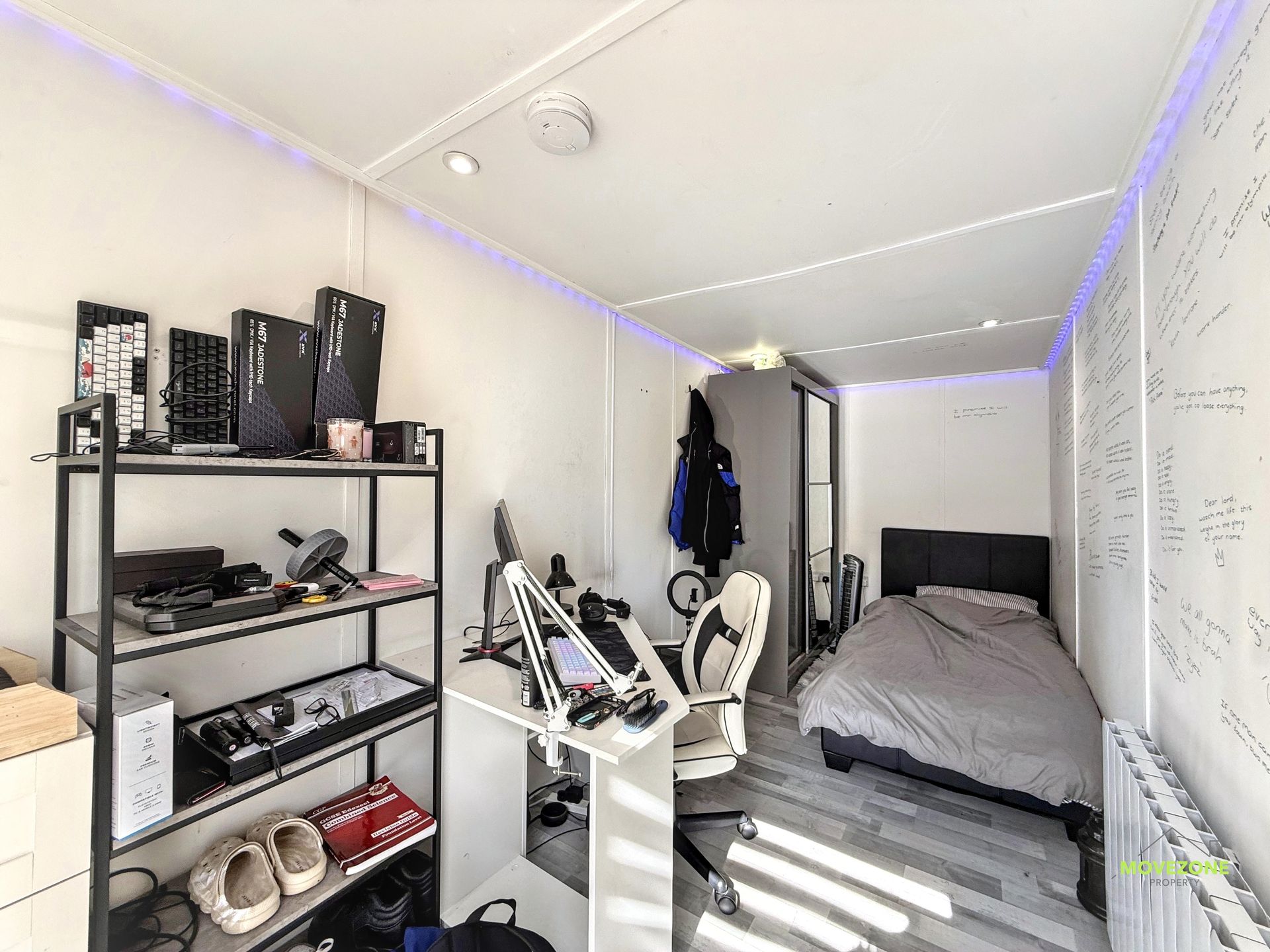 Small bedroom with bed, shelves, exercise equipment, and wall-mounted whiteboards under blue LED lights.