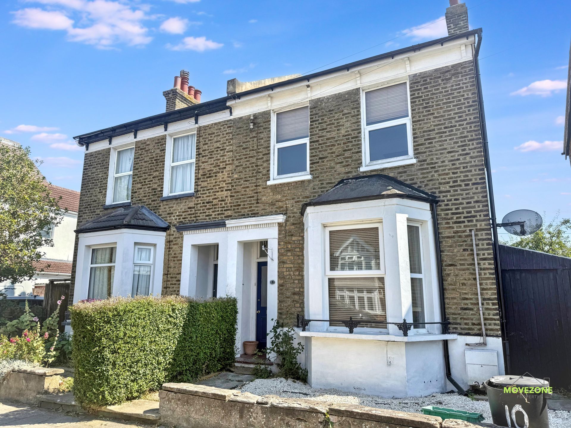 Semi-detached brick house with white trim and a small front hedge on a sunny day