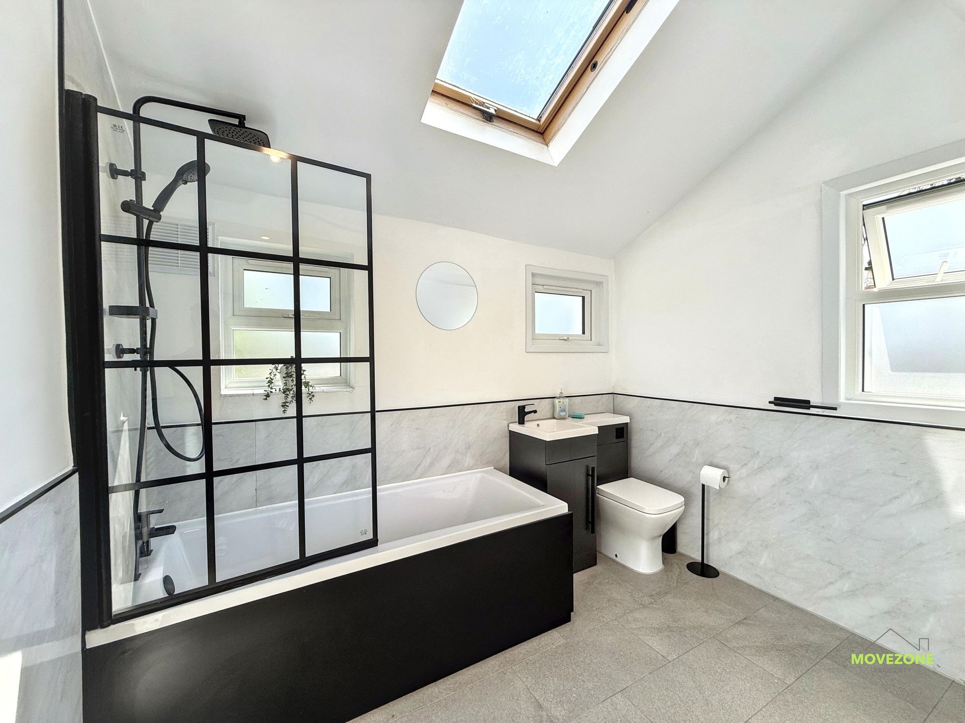 Modern bathroom with black-framed shower, freestanding tub, toilet, vanity, and skylight