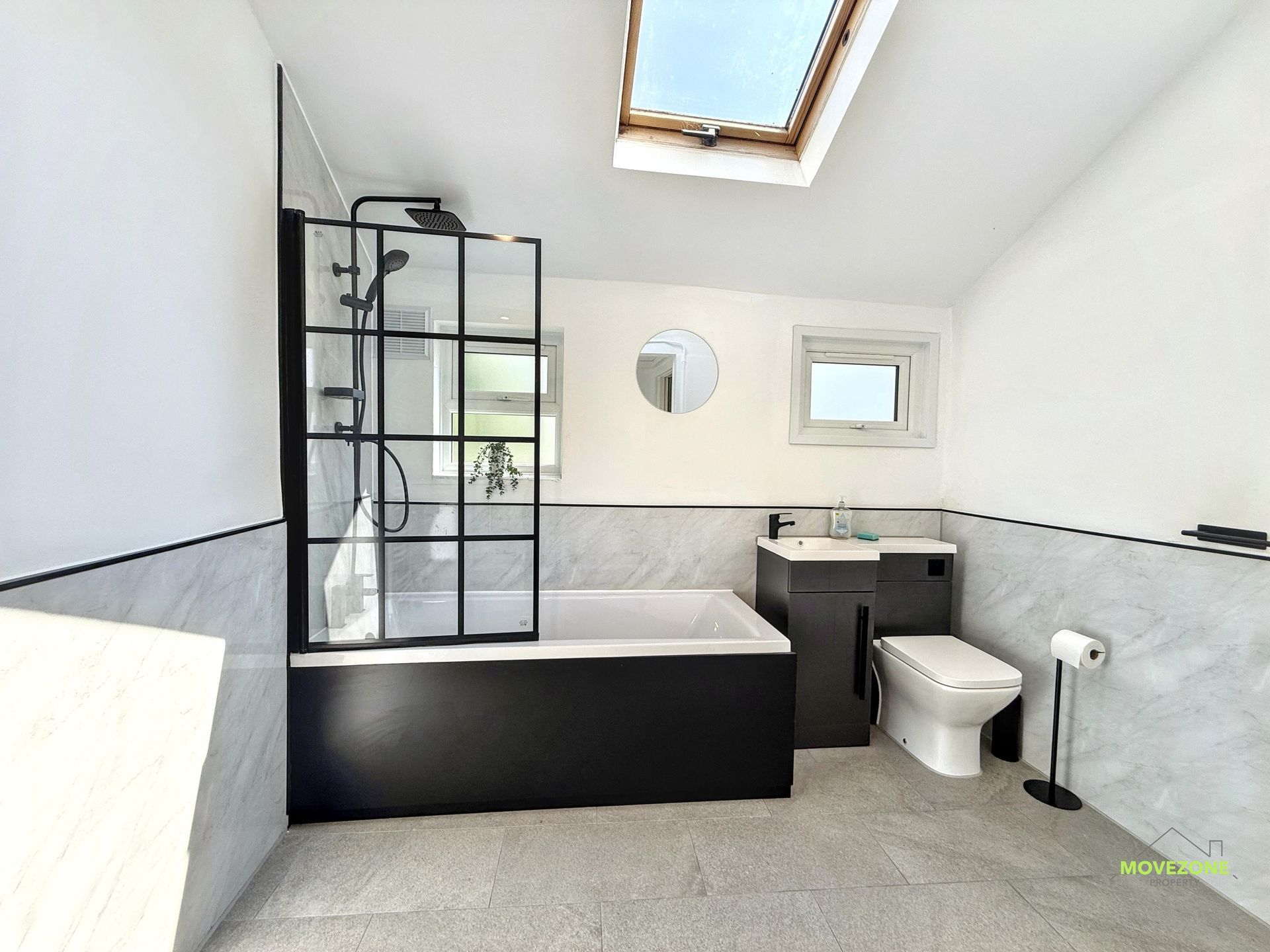 Modern bathroom with black tub, glass shower, skylight, sink, toilet, and gray tiled walls