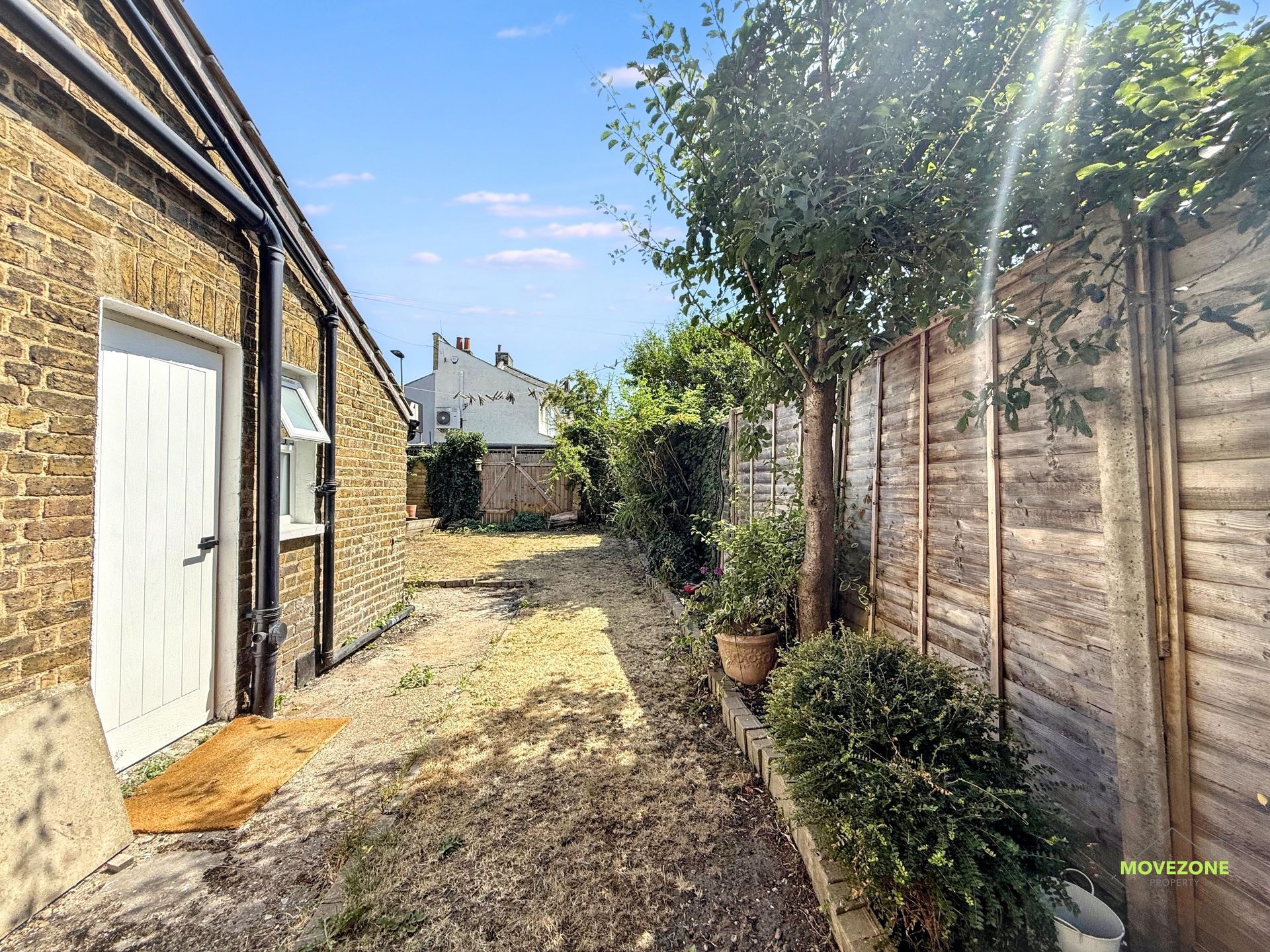 Narrow sunny backyard walkway beside brick and wood walls, with doors, gravel path, and greenery.