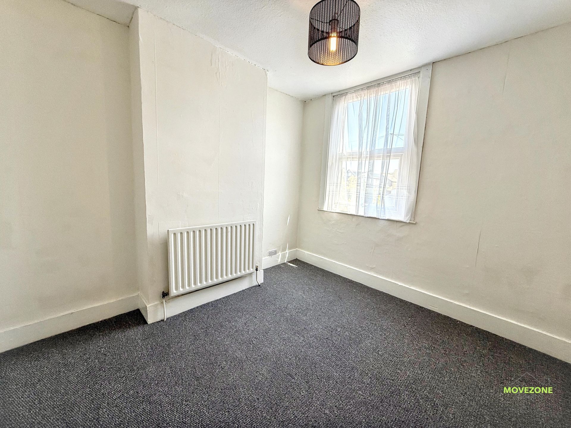 Empty white room with gray carpet, radiator, window, and a ceiling light.