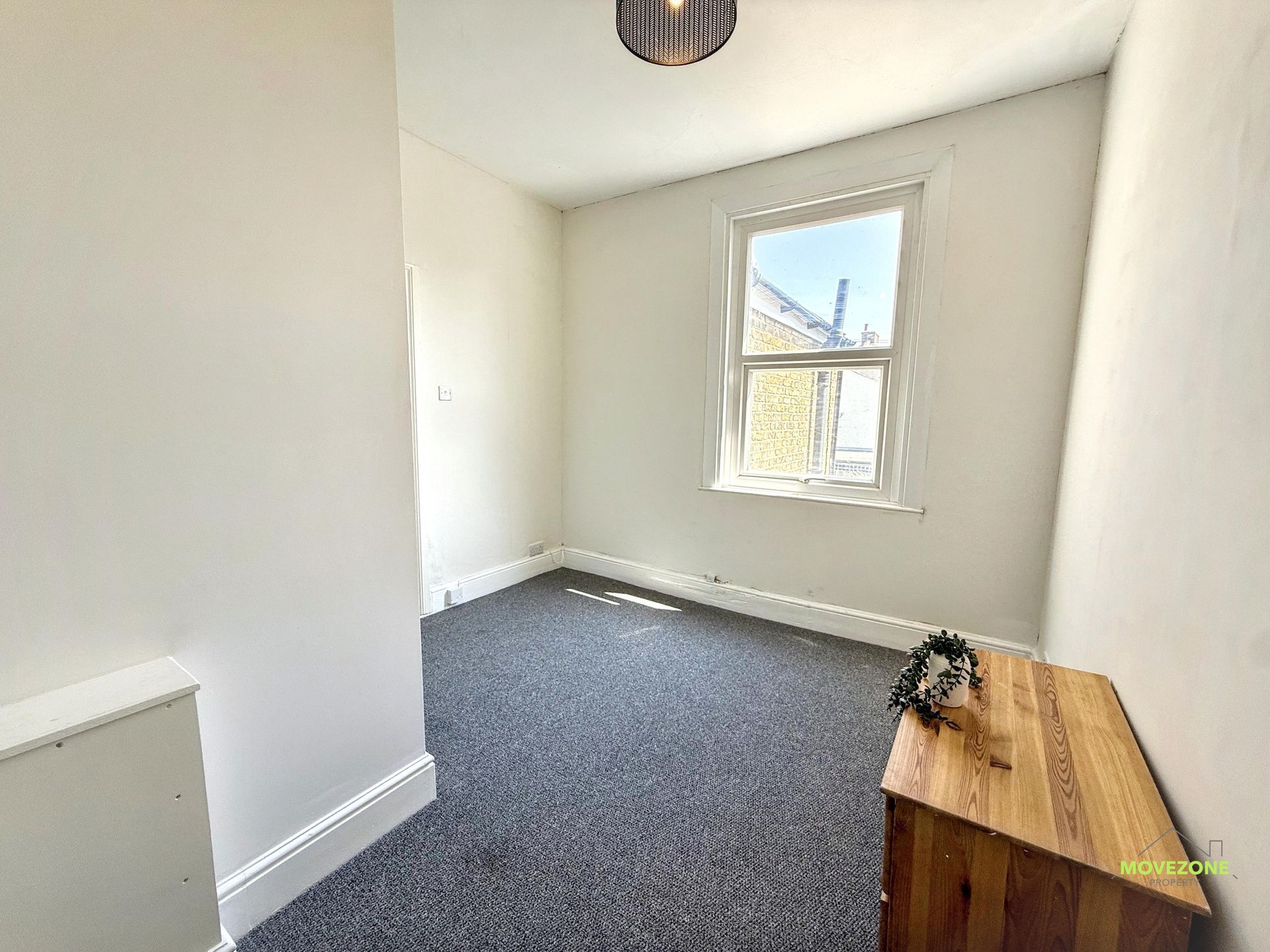 Empty white room with gray carpet, a window, and a small wooden bench with a plant.