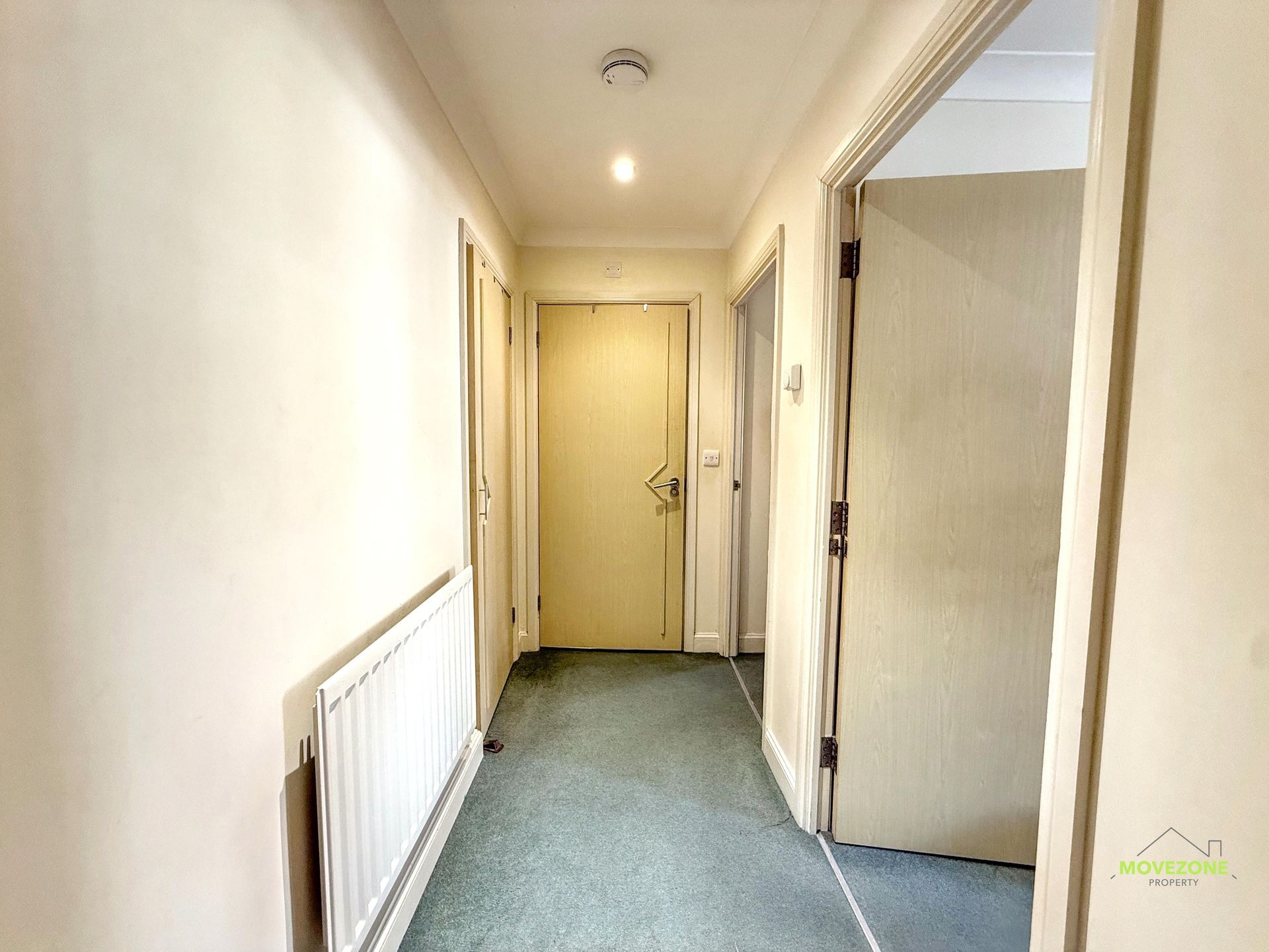 Narrow hallway with cream walls, green carpet, radiator on left, and doors at the end and right