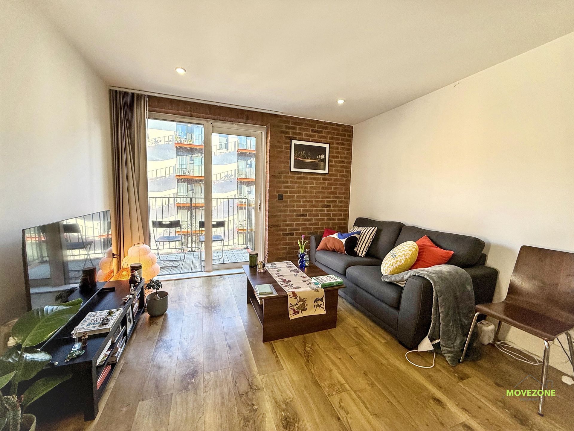 Bright living room with couch, TV, coffee table, brick accent wall, and balcony doors facing city buildings