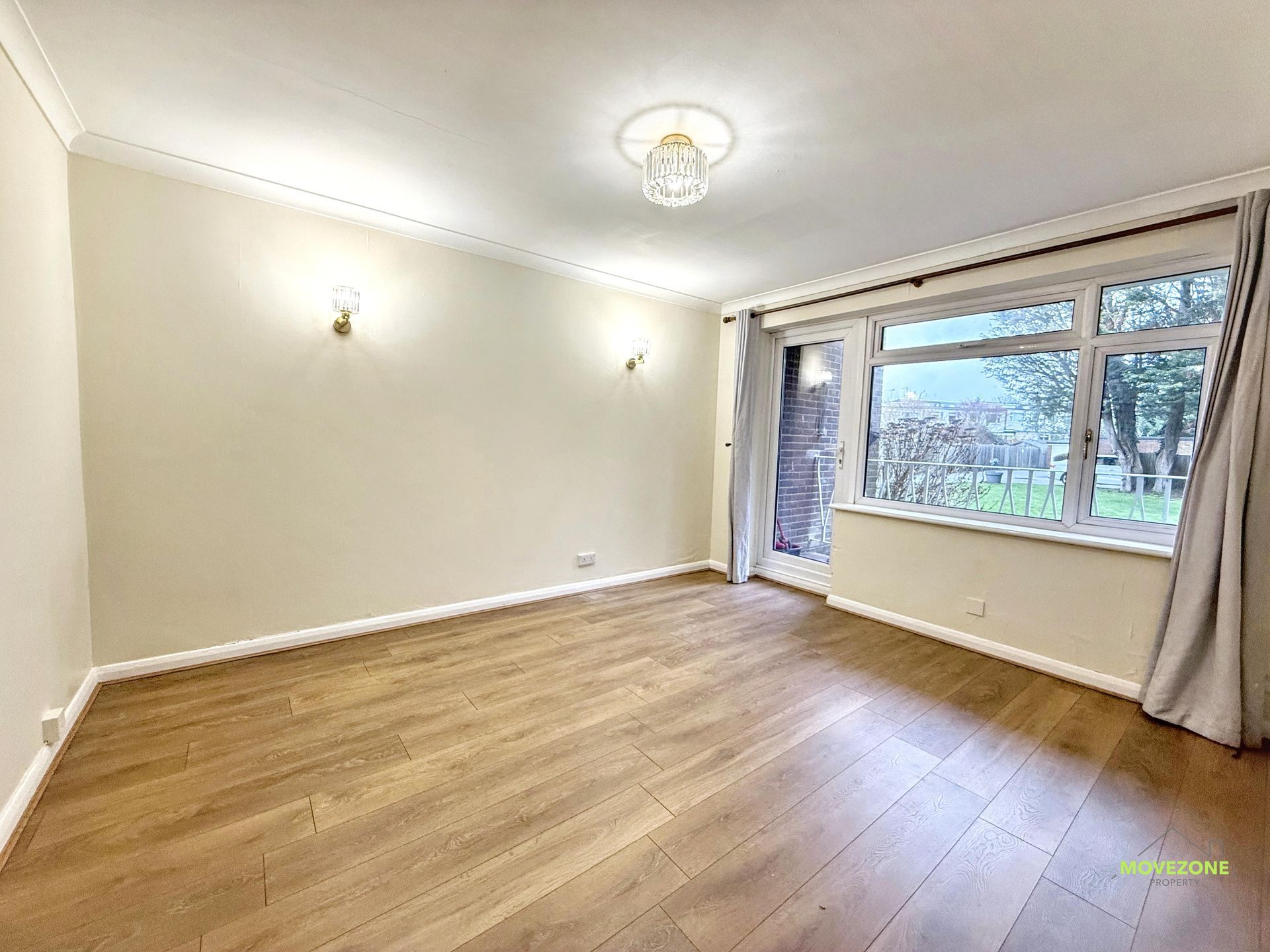 Empty unfurnished living room with wood floor, cream walls, and large window overlooking trees.