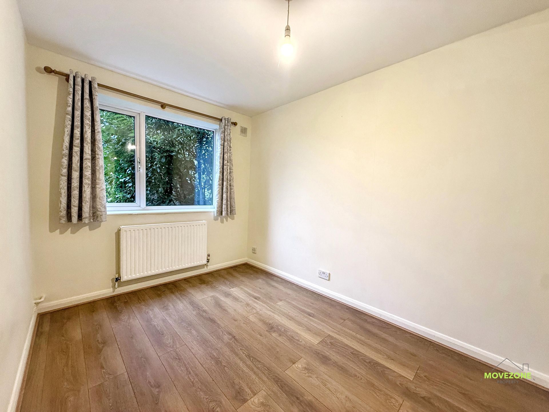 Empty beige bedroom with wood floor, window, radiator, and a hanging light fixture.